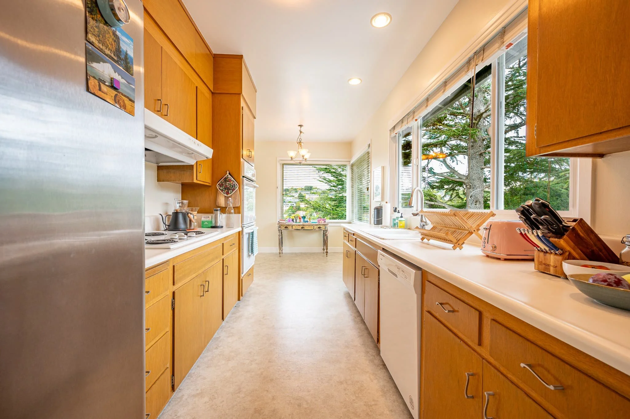 A kitchen with wooden cabinets, a white countertop, and large windows with a view of trees outside. There are various kitchen appliances and utensils on the counter, including a toaster and a dish drying rack.