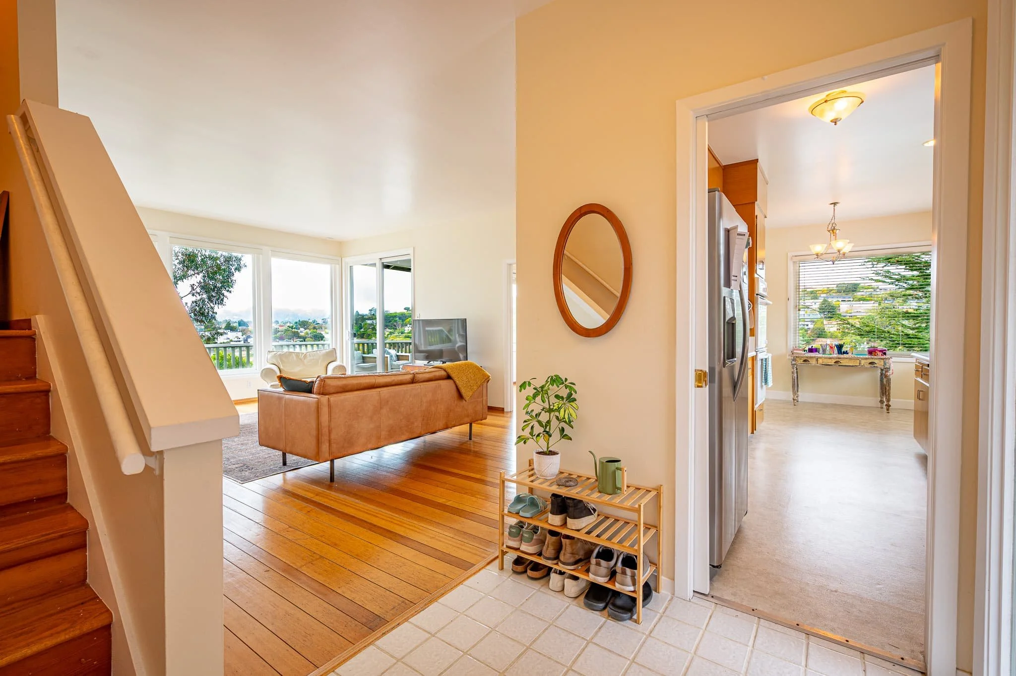 View of a cozy living room with large windows and a balcony, from an entryway with a shoe rack and tiled floor.