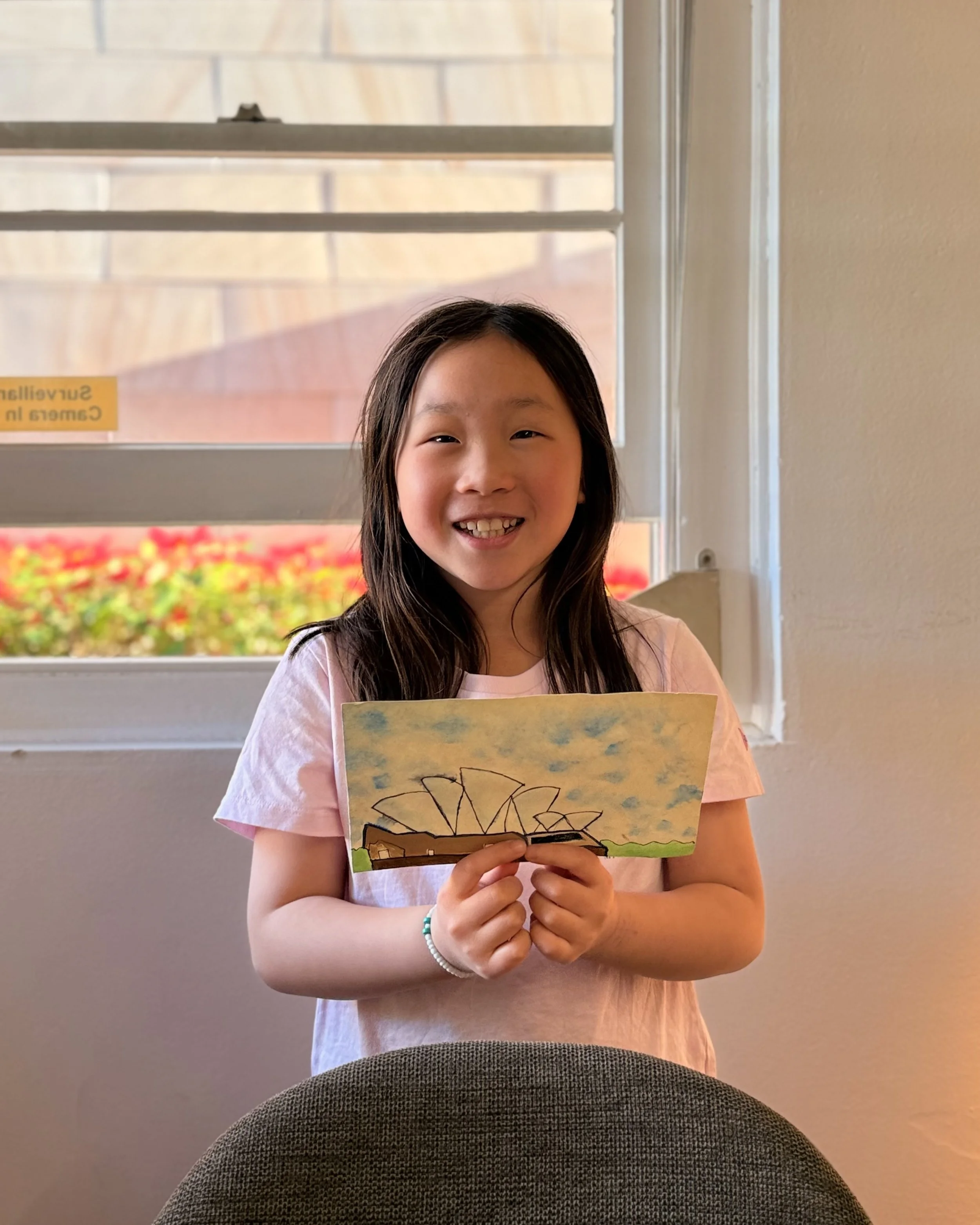 A young girl with long dark hair is smiling and holding a drawing of the Sydney Opera House, standing indoors near a window.