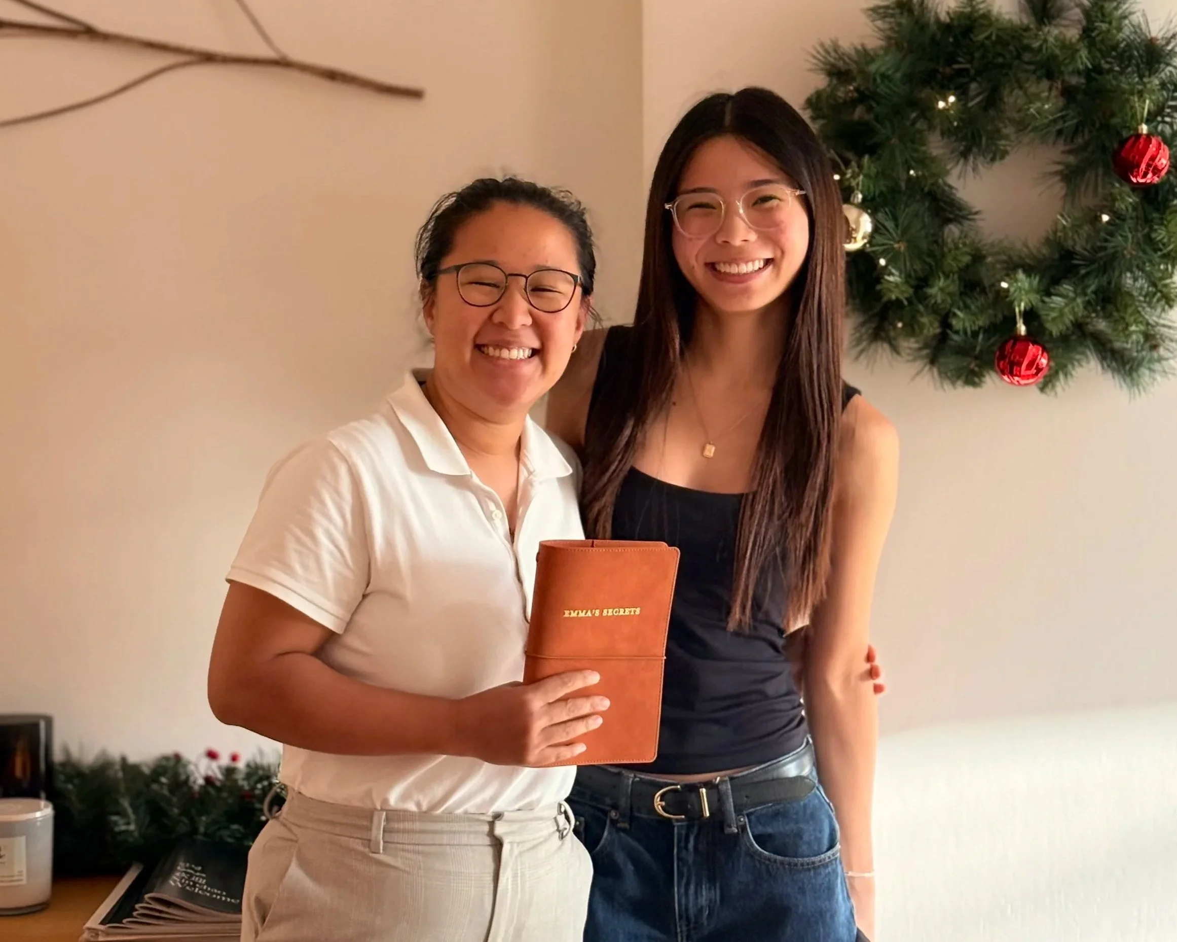 Two women smiling for a photo near a decorated Christmas tree, one holding a journal that says 'Emma's Secrets' on the cover.