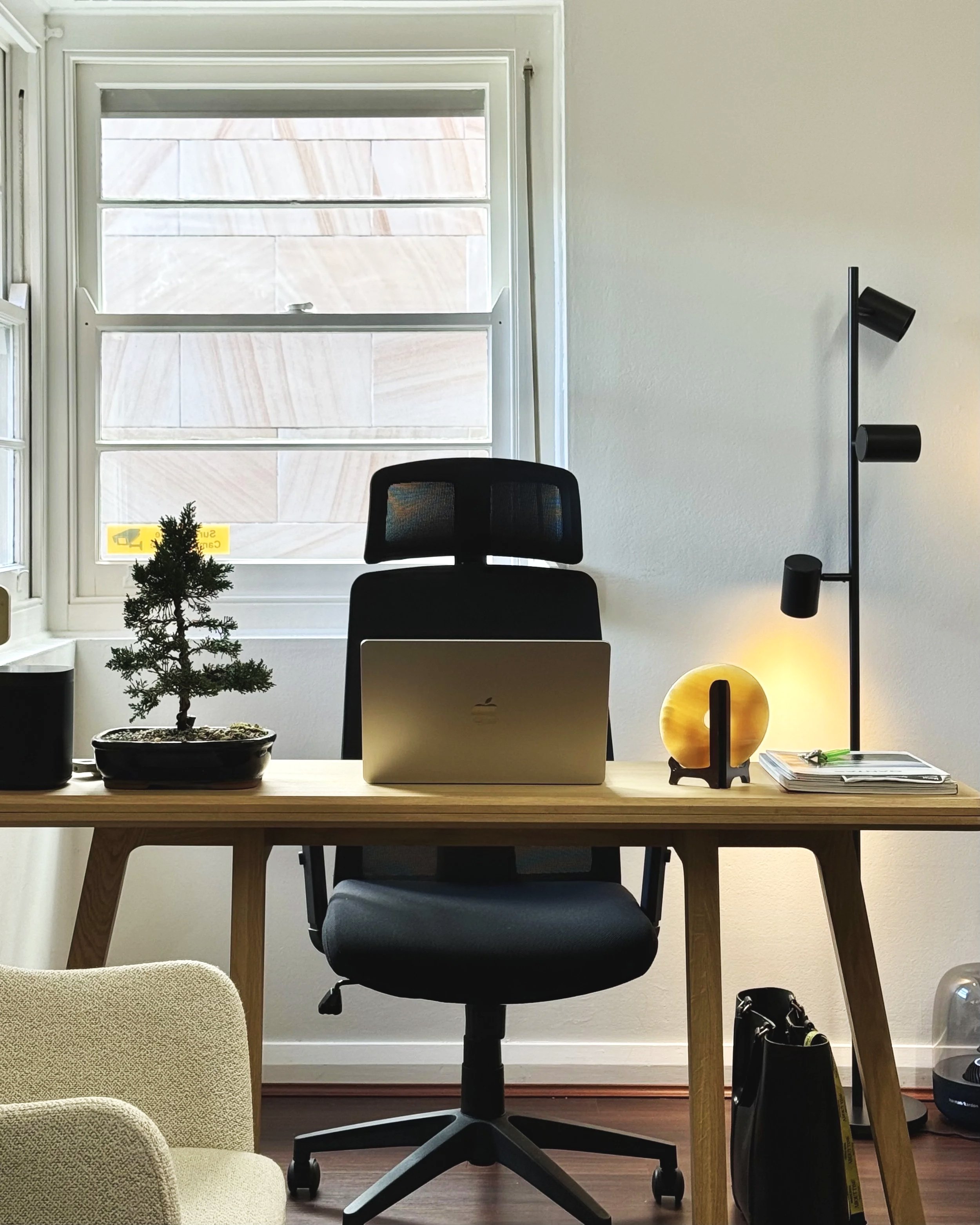 Modern home office with a wooden desk, black ergonomic chair, a small potted tree, a yellow circular lamp, a laptop, and a tall black floor lamp with three adjustable lights. A window with blinds is behind the desk, letting natural light in.