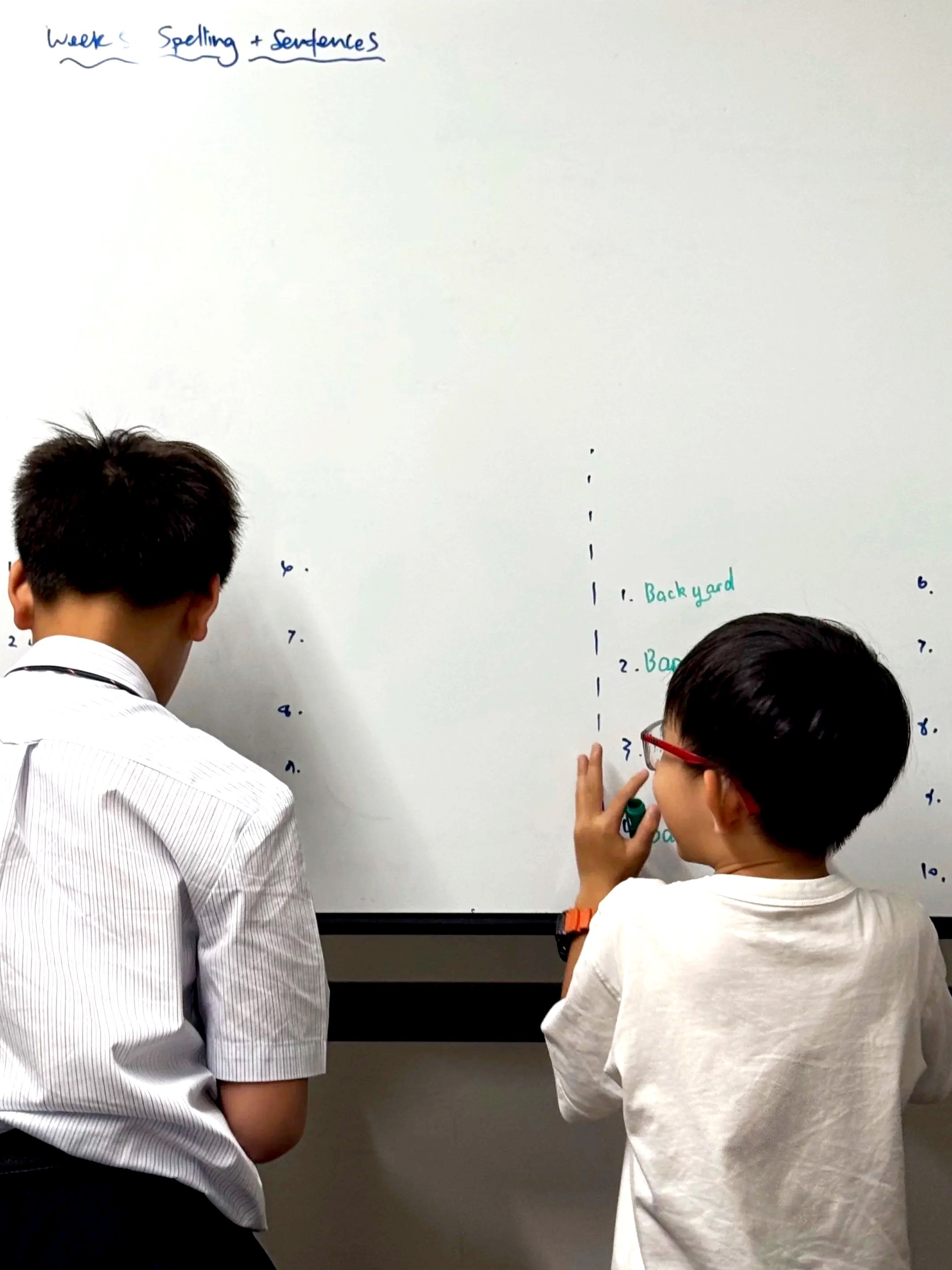 Two boys standing in front of a whiteboard, working together on a list. The board has text written in different colors, including 'Week's Spelling + Sentences' at the top and numbered items underneath, with some words like 'Backyard' and 'Back' visible.