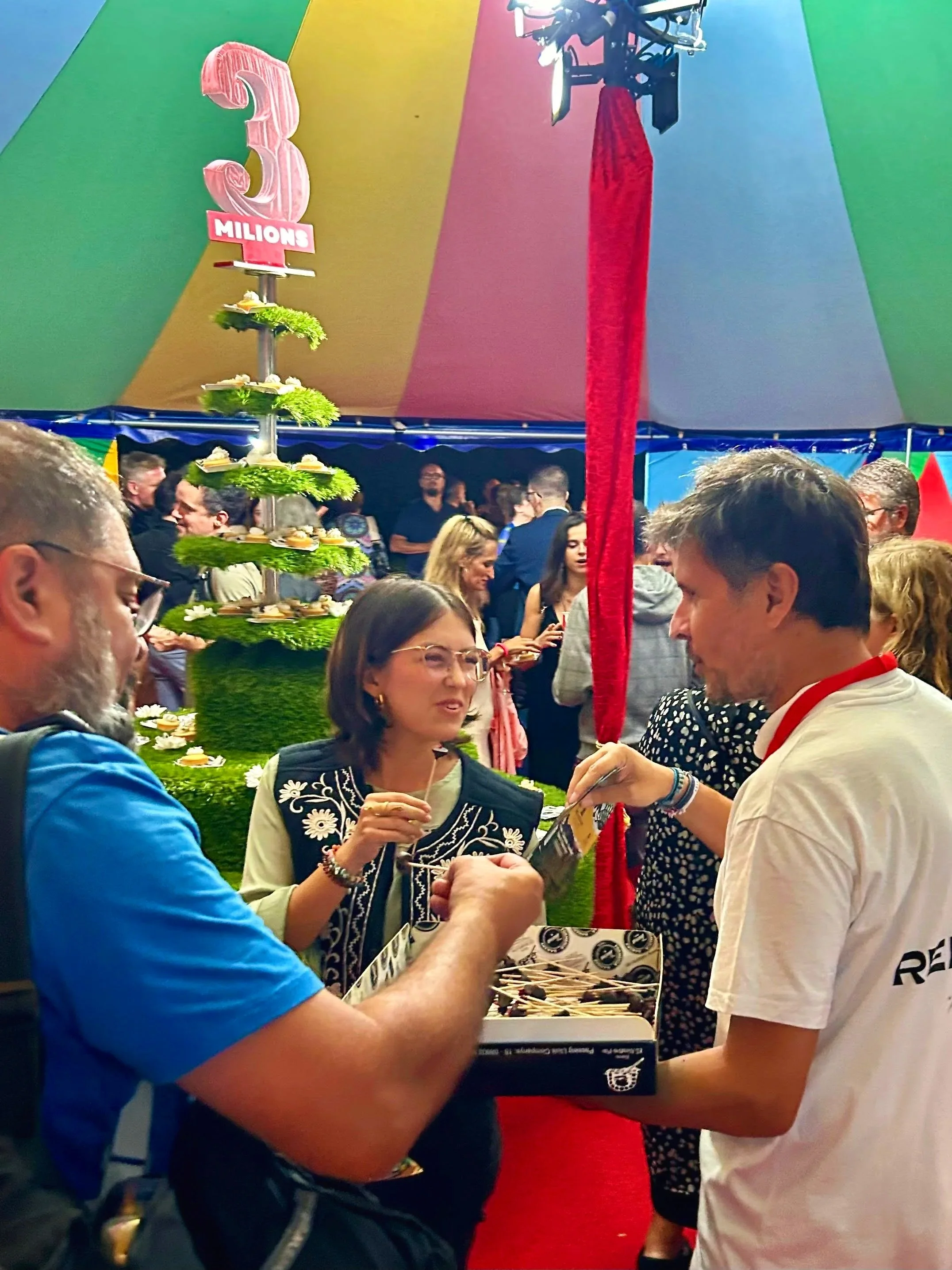 Personas interactuando en un evento con un gran pastel decorativo en forma de número 3 millones en un ambiente colorido