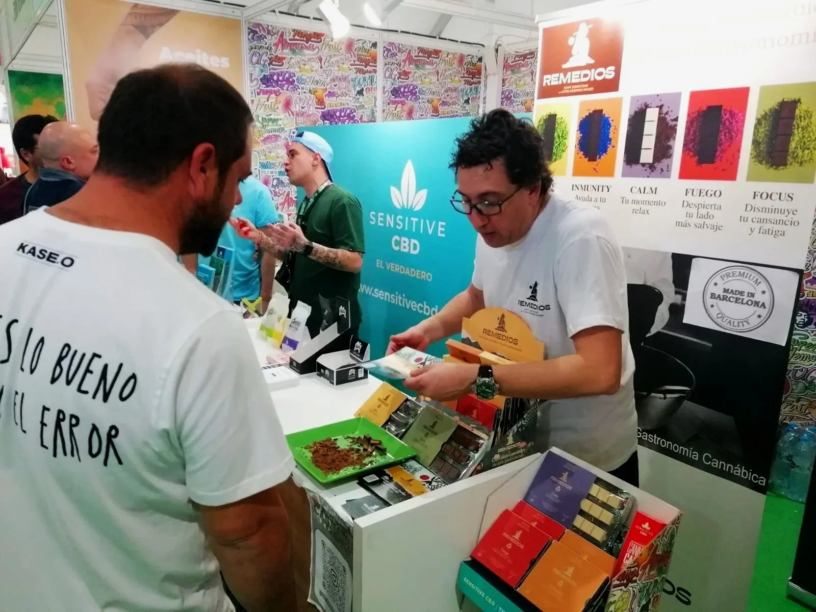 Persona en un stand de exhibición revisando productos de CBD, con otros visitantes y representantes en el fondo.