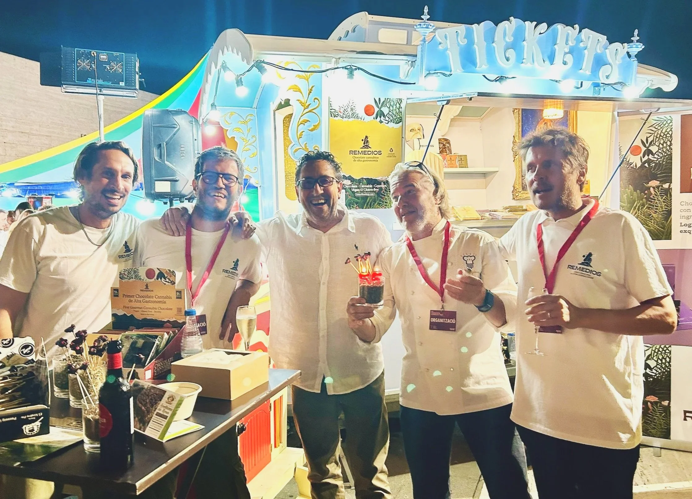 Un grupo de cinco hombres sonrientes en una feria, con un stand de chocolates y productos de cannabis en el fondo, algunos con medallas y uno sosteniendo un pastel pequeño con antorchas.