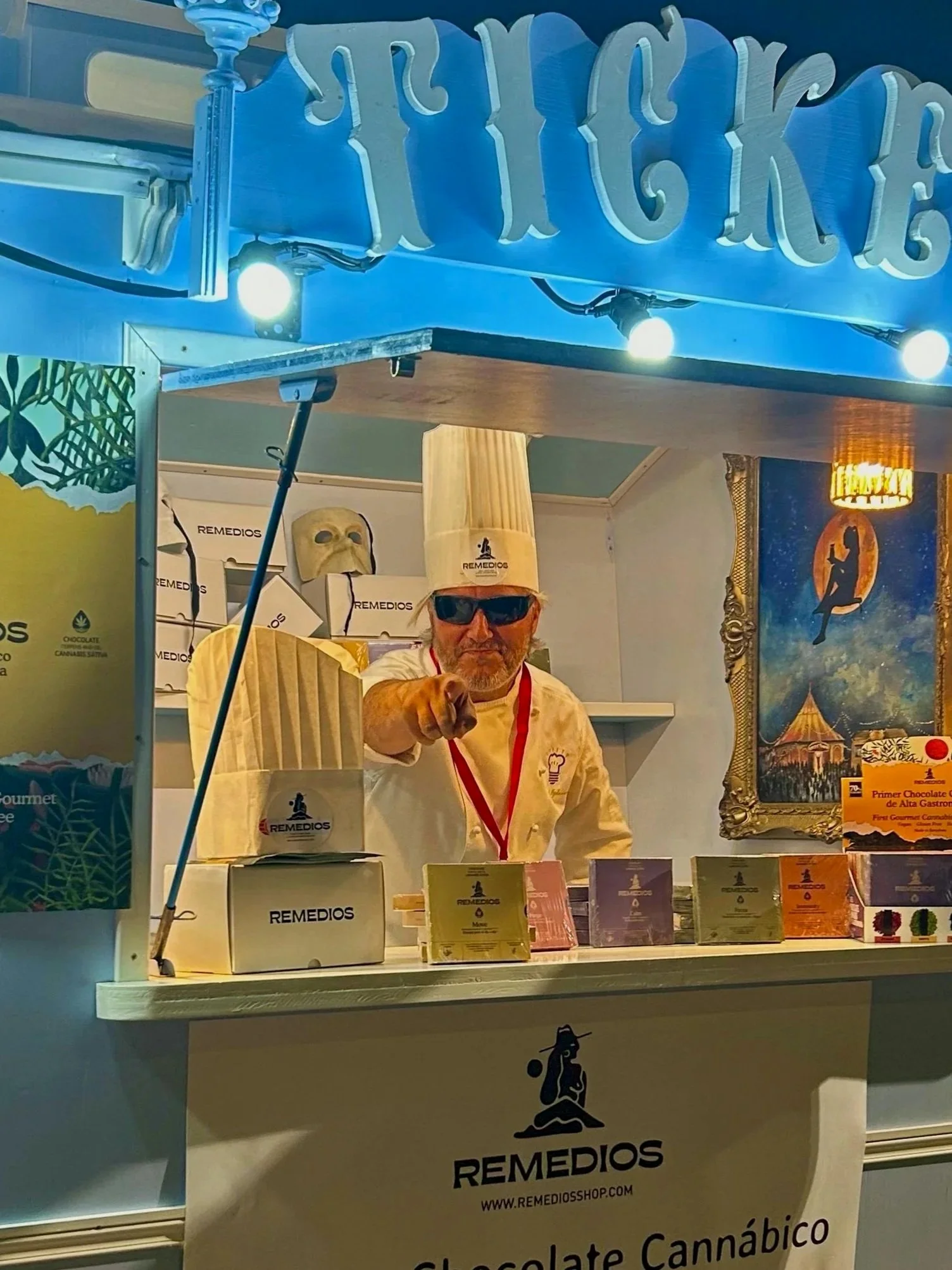 Hombre con barba, gafas oscuras y gorro de chef señalando hacia la cámara desde un puesto de venta de chocolates en un evento. Presenta productos de chocolate en cajas y carteles en un stand colorido con declaración de marca REMEDIOS.