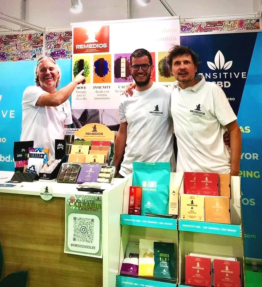 Tres hombres en una feria o exposición promocionando productos de la marca Remedios, con mesas que tienen cajas y paquetes de productos y un cartel detrás con información sobre la salud y el bienestar.