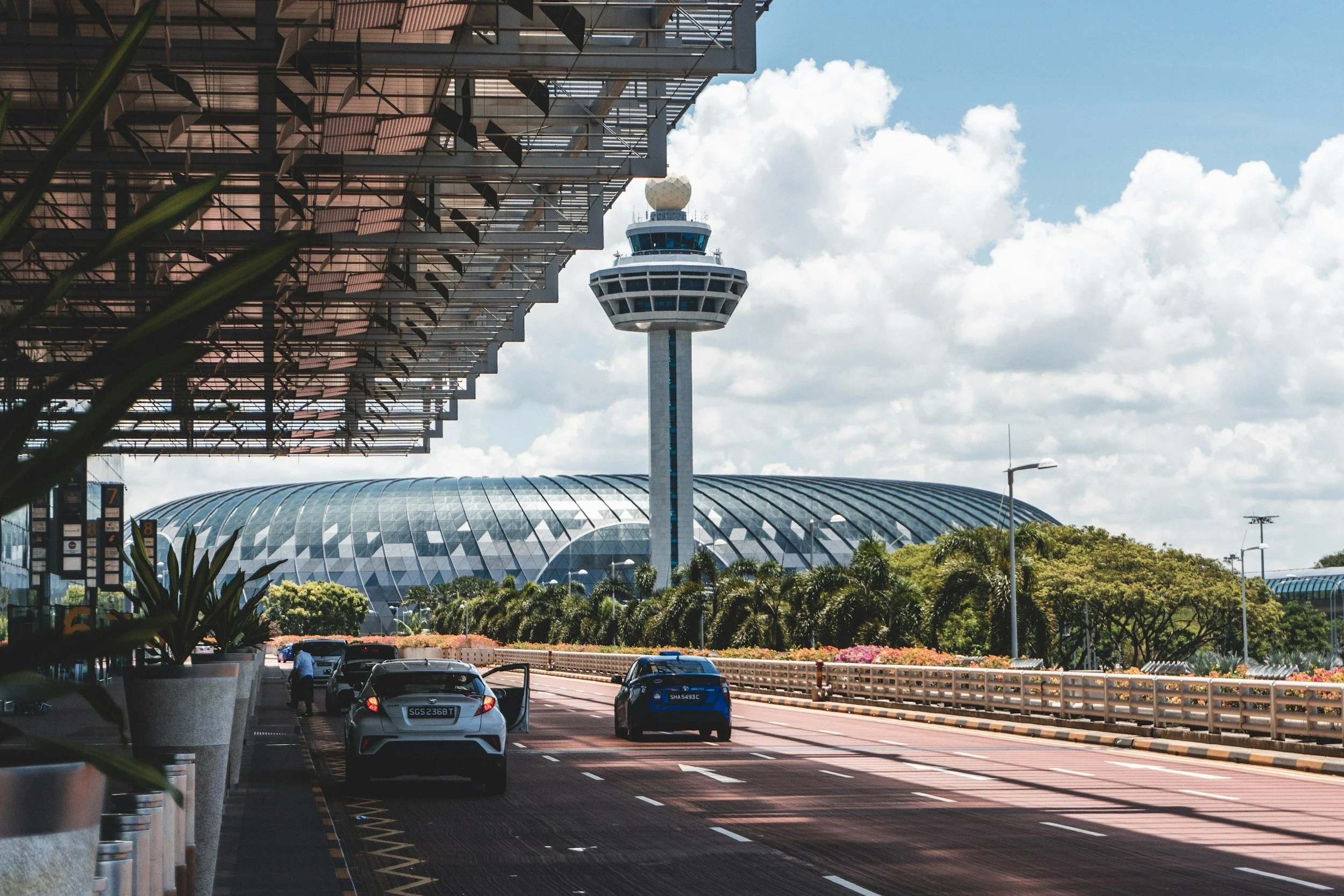 View of an airport terminal with a control tower in the background, cars parked and walking along the street, and lush green trees under a partly cloudy sky.