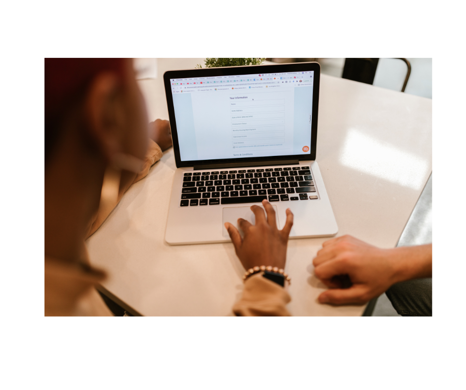 Person filling out an online form on a laptop, with another person sitting beside them and their hand on the table.