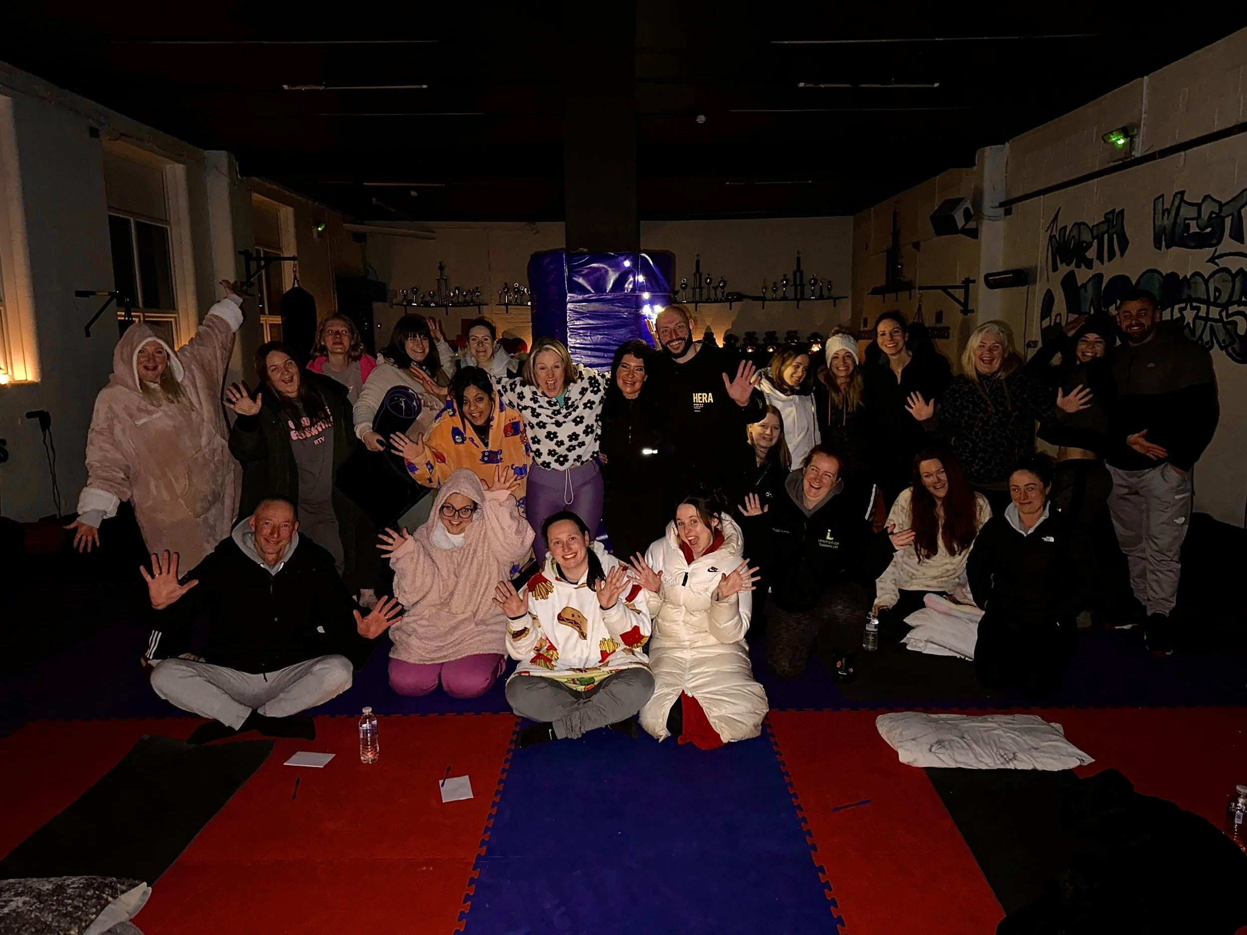 Group of people in pajamas and comfy clothes, smiling and waving, seated and standing on a padded floor in a dimly lit room with dark walls and some graffiti, celebrating or having fun together.