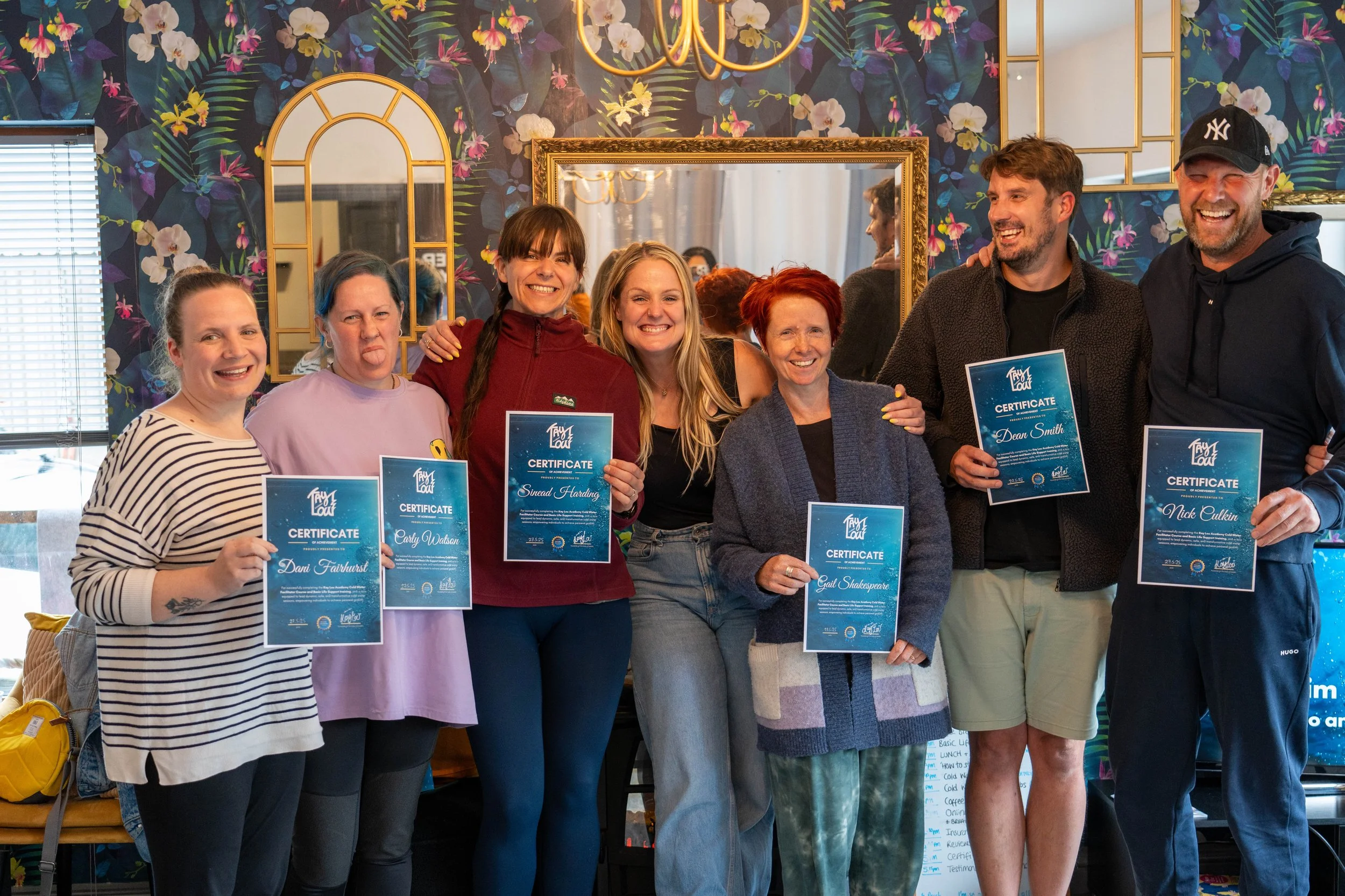 Group of seven people smiling and holding certificates, standing in front of a decorative floral wallpaper with a gold-framed mirror. The group includes five women and two men, dressed casually, celebrating an achievement.