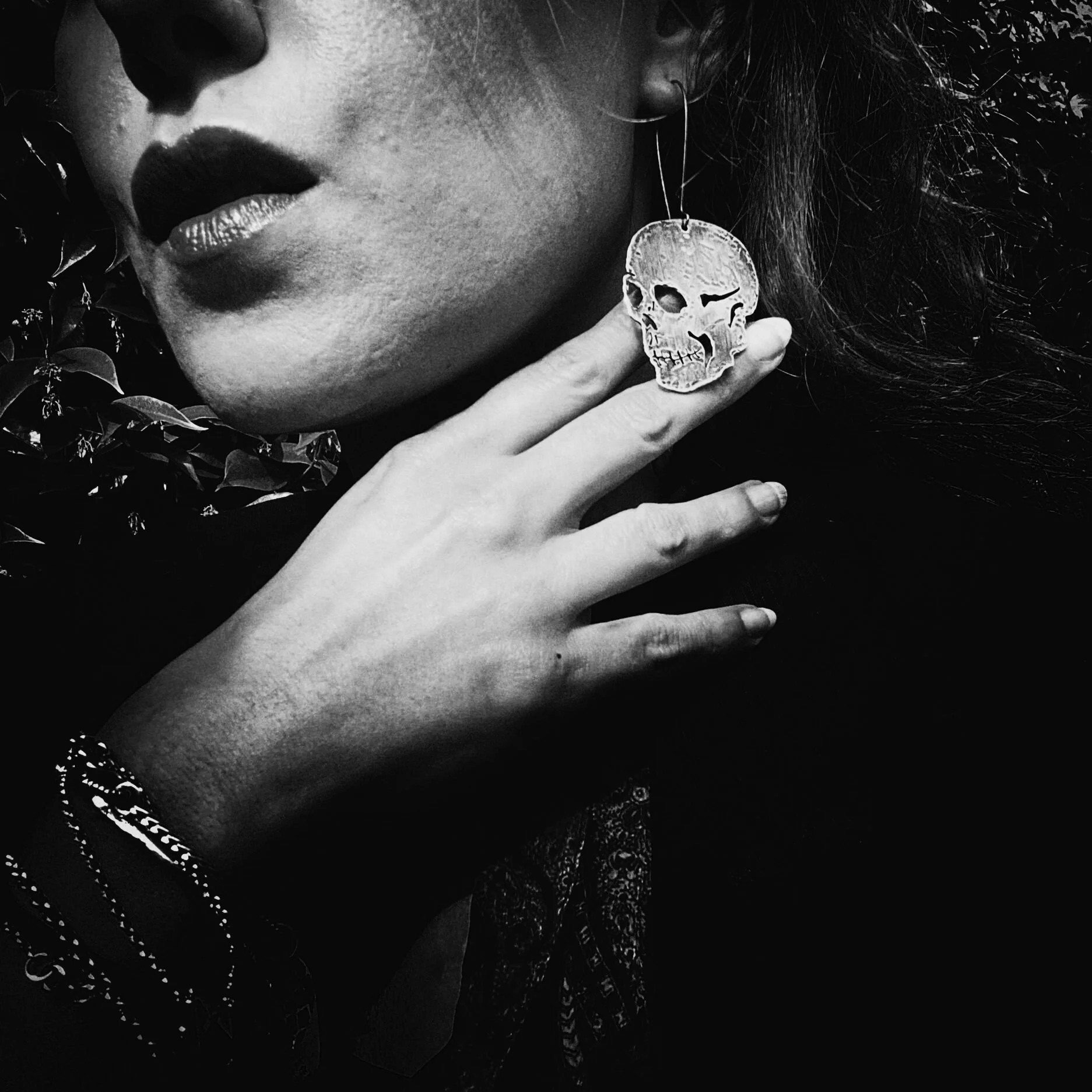 A woman with dark lipstick, wearing large earrings shaped like a skull and bracelets, touching her neck with her hand in a close-up black and white photo.