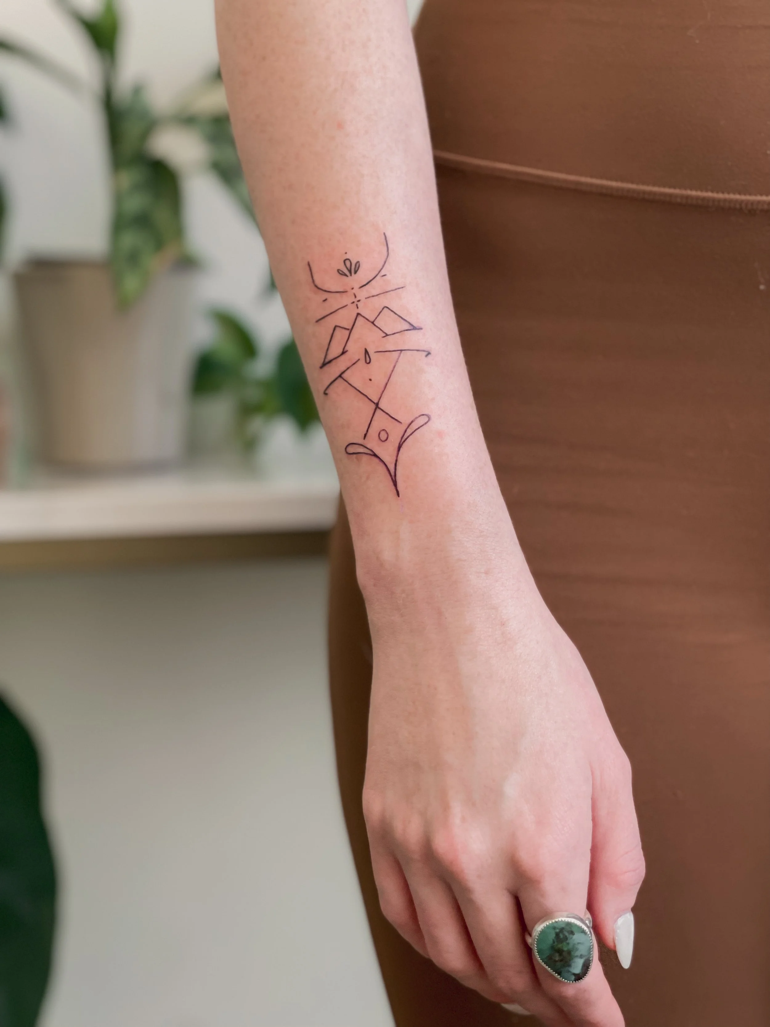 A person has a tattoo on their forearm of an abstract, minimalist line drawing of a woman sitting with legs crossed and hands on knees, with stylized facial features and a small plant sprouting from the head.