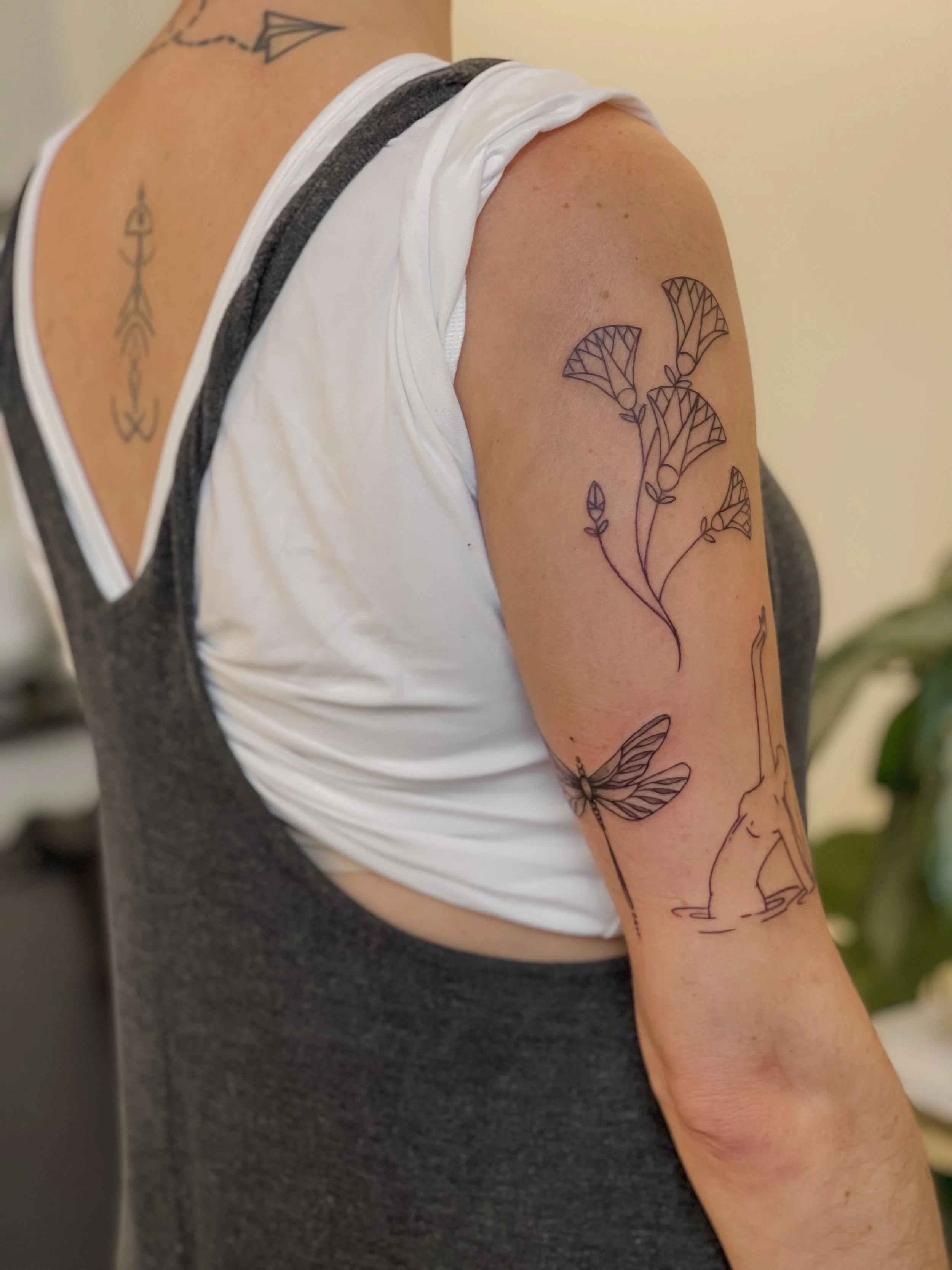 Close-up of a person's upper arm and shoulder with multiple black ink tattoos, including a bee, ginkgo leaves, and a minimalistic arrow, wearing a white tank top layered over a gray shirt.