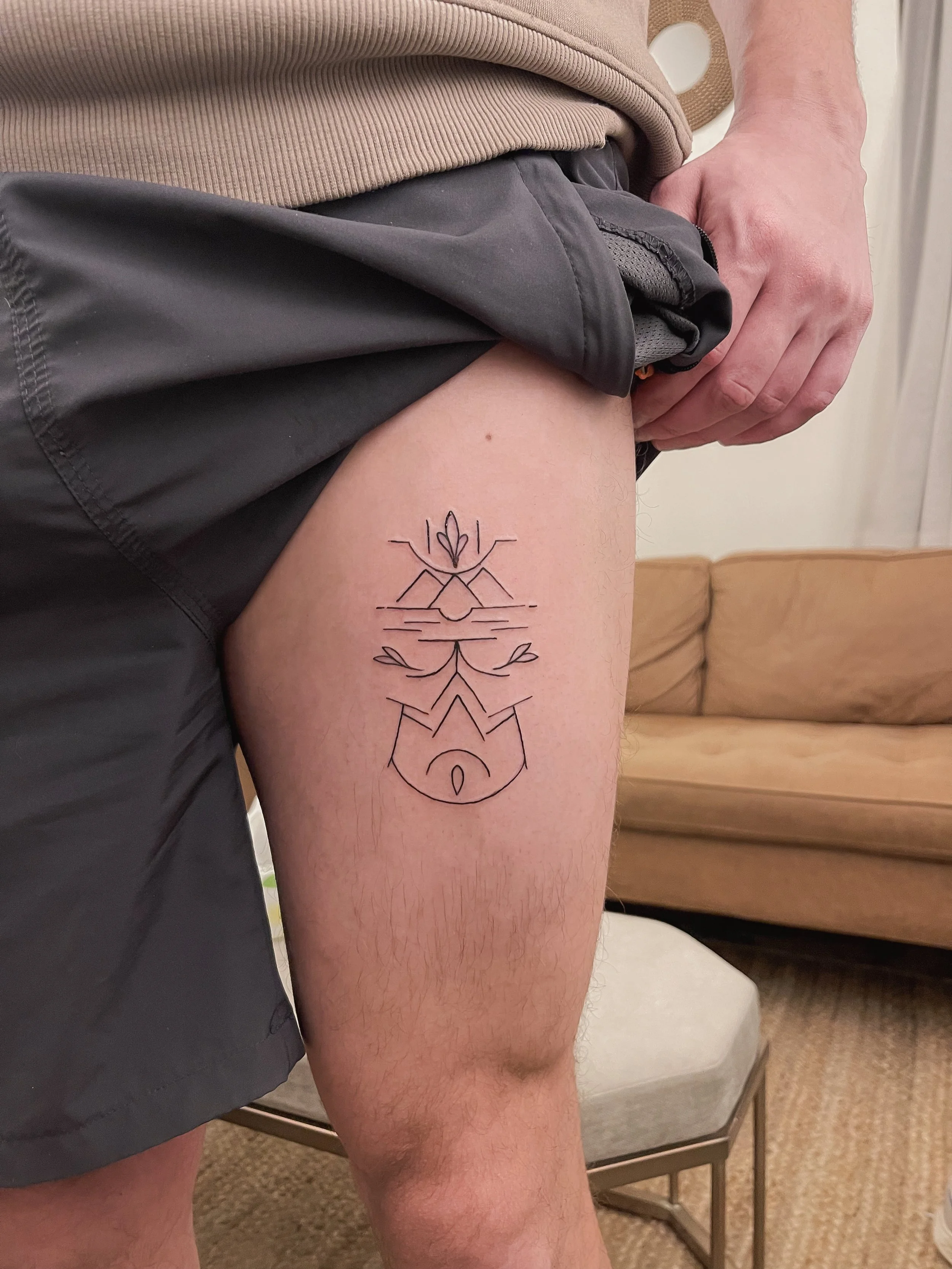 A person's upper thigh with a black line tattoo featuring abstract geometric and natural elements, including a flower, lines, and shapes.