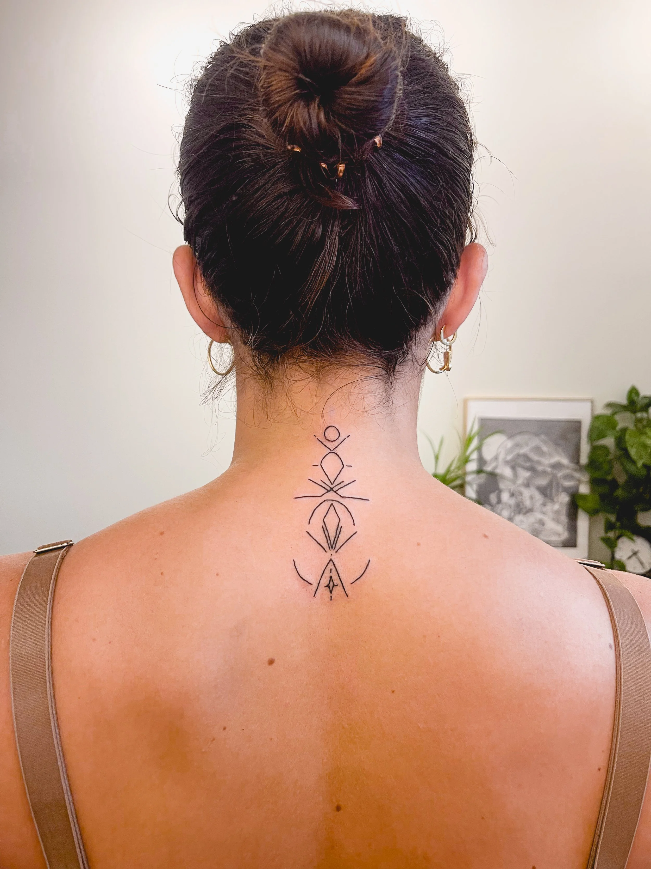 Back of a woman with dark hair styled in a bun, showing a geometric tattoo on her upper back and neck, with a framed black and white artwork and greenery in the background.