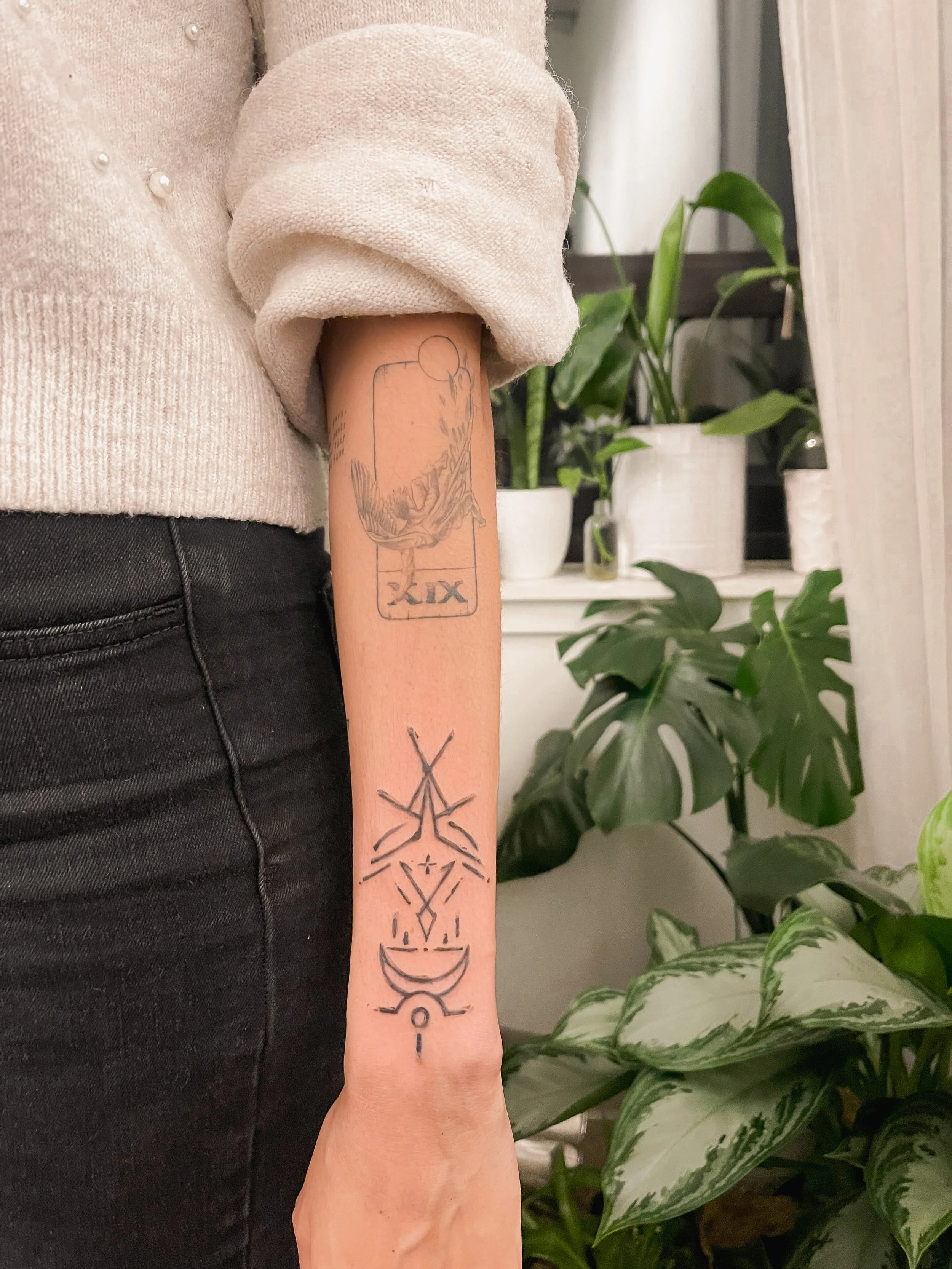 A person's forearm with two tattoos: one of a tarot card that appears to be the ten of swords, and another of a geometric, mystical design with abstract shapes and lines, including a crescent moon.