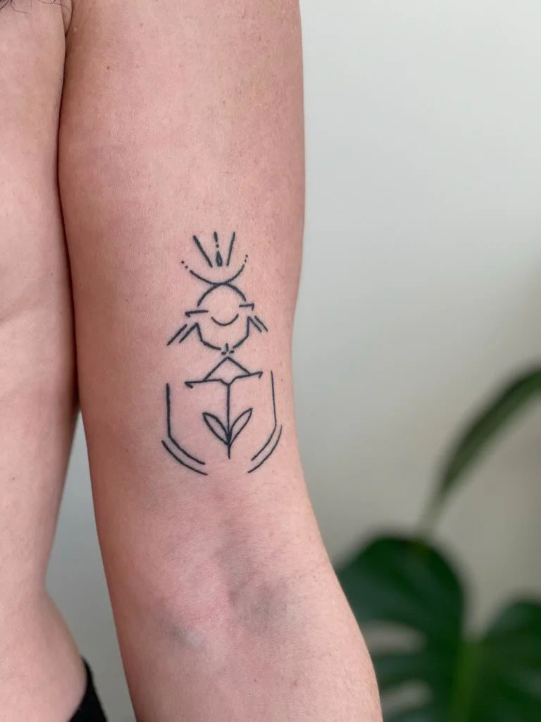 A minimalist black line tattoo on a person's inner forearm that features a symmetrical design combining abstract shapes, a small plant with leaves, and decorative elements.