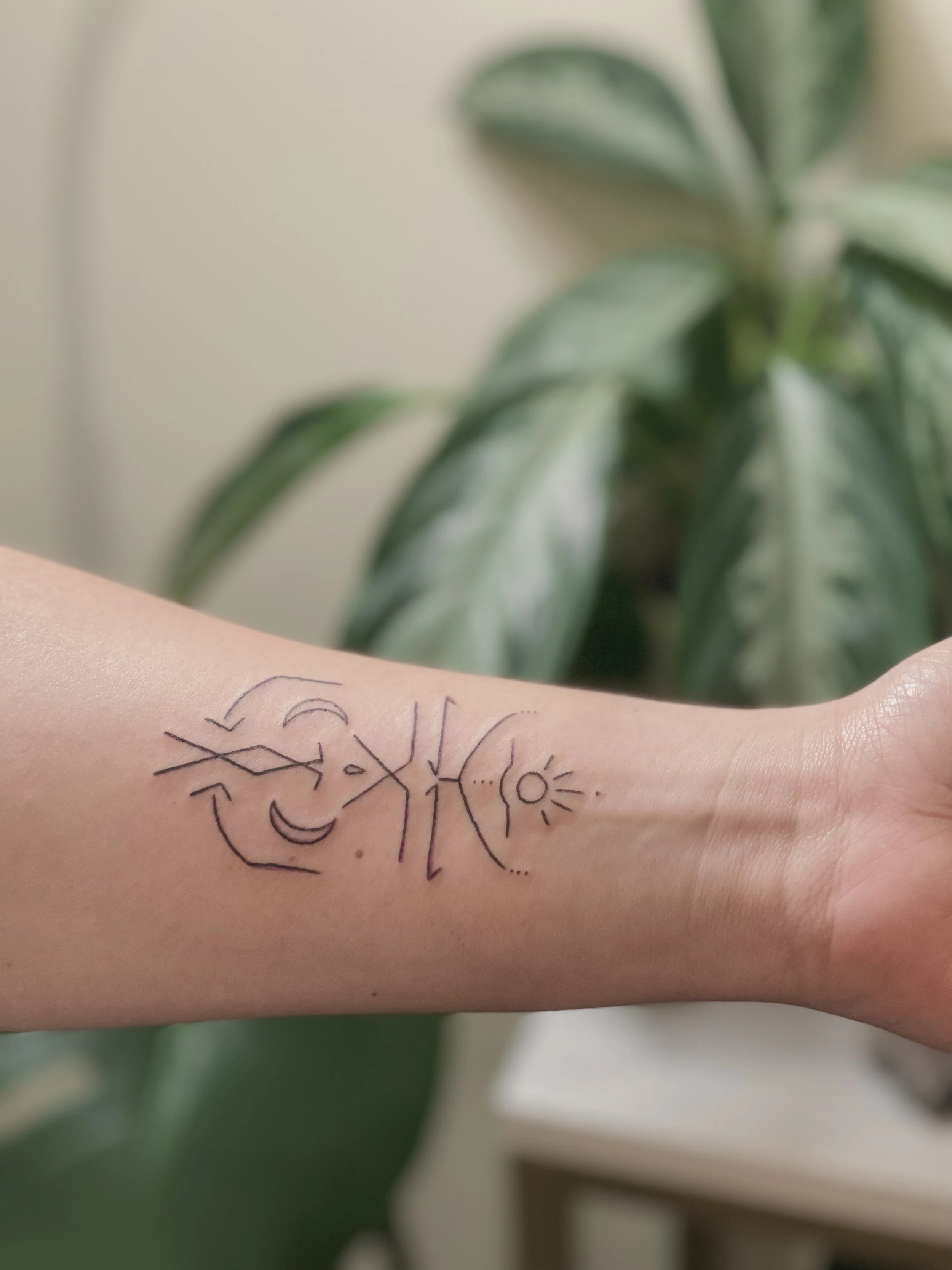 Tattoo of abstract black line art on inner forearm with design resembling a face, moon, and sun symbols, with green leafy plant in blurred background.