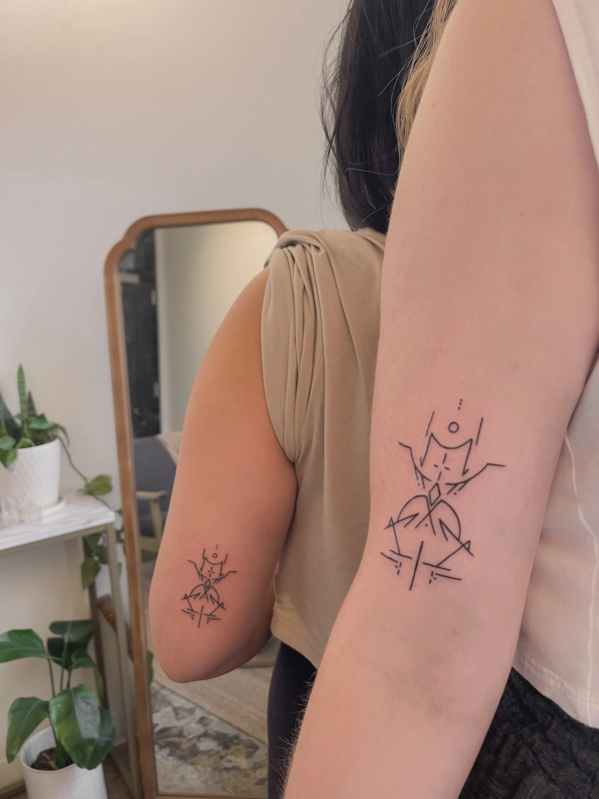 Two tattoos of abstract, geometric designs on the upper arms of a person. The tattoos feature lines and shapes resembling stylized anatomical figures.