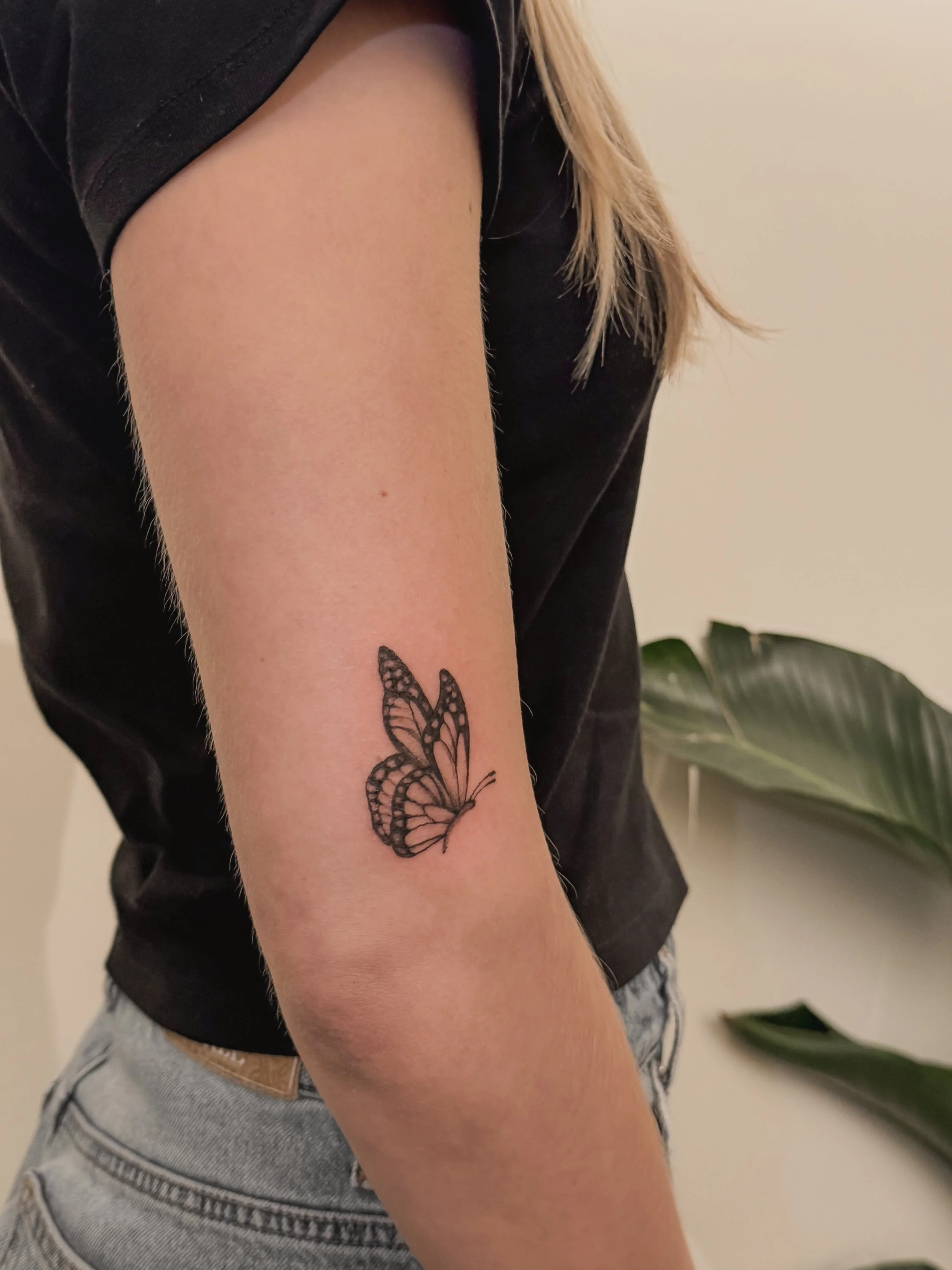 A tattoo of a butterfly on a person's upper arm, with a black shirt and light-colored jeans.