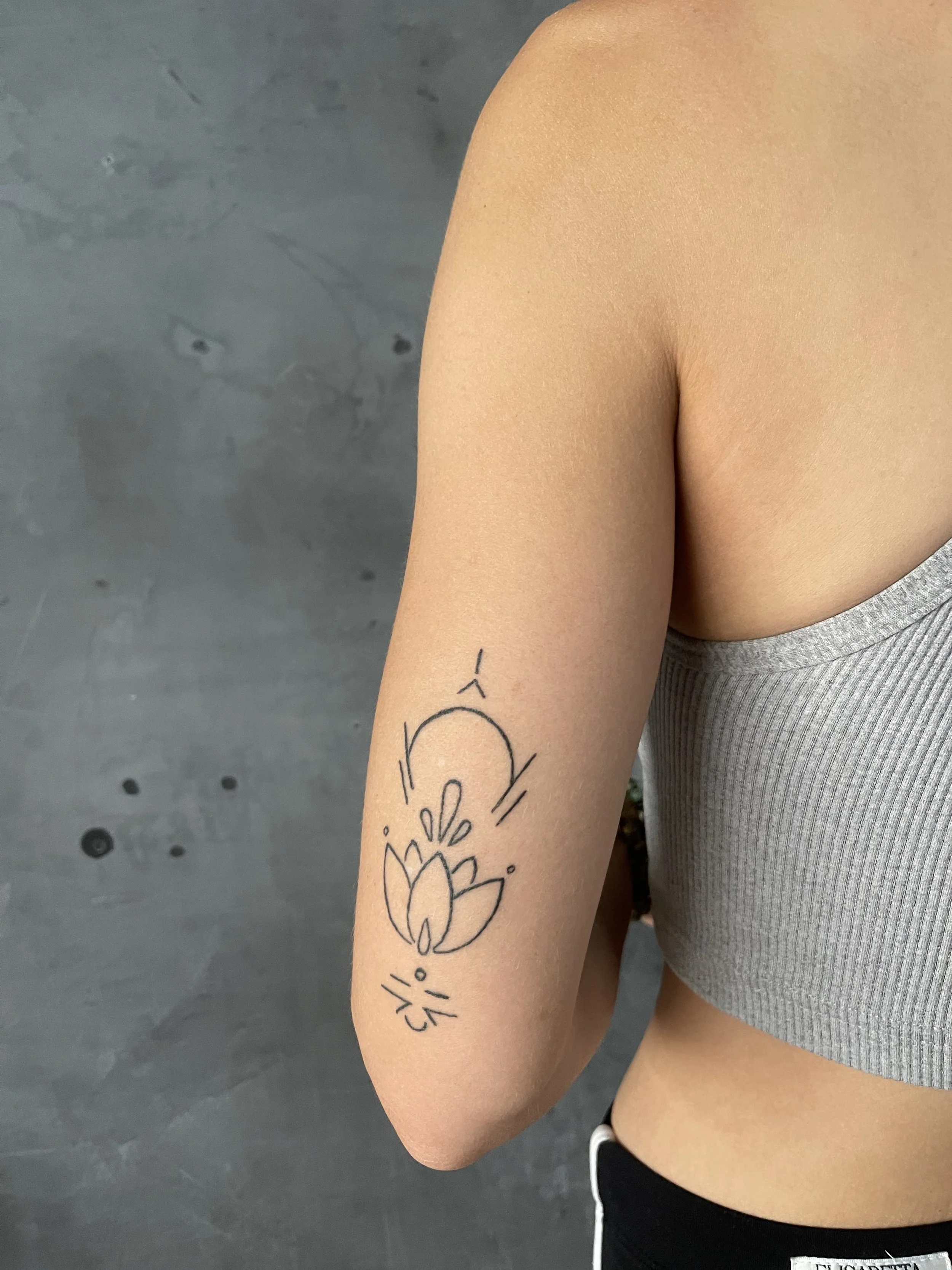 A close-up of a person's upper arm with a tattoo of a stylized lotus flower and abstract lines, wearing a light gray sleeveless top, against a gray background.