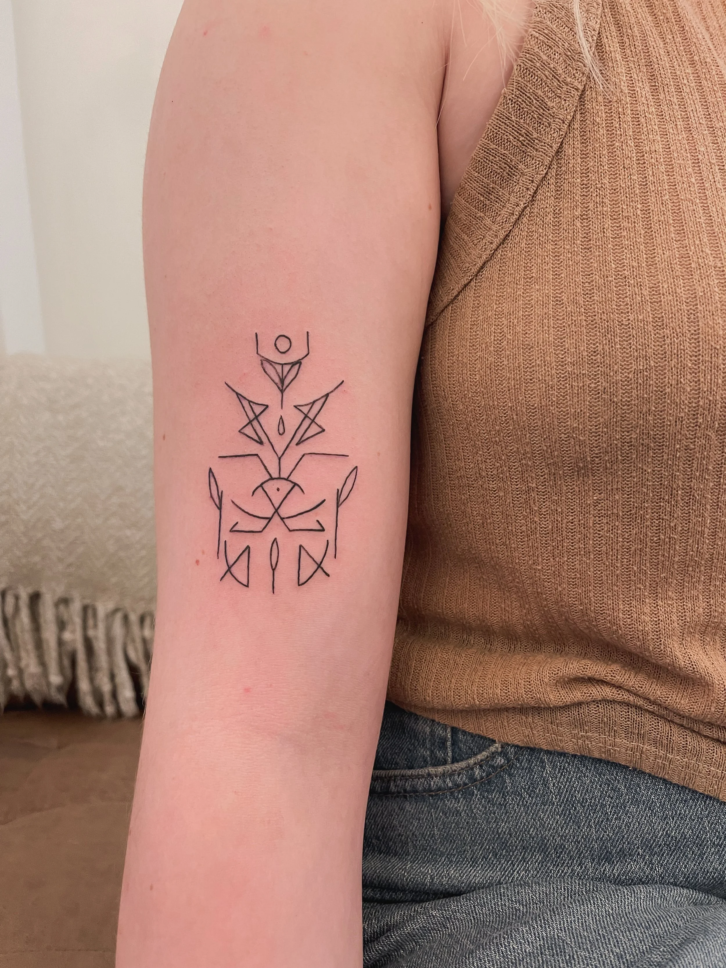 A person's upper arm with a black geometric tattoo design, wearing a brown sleeveless top and blue jeans, and a cream-colored textured blanket in the background.
