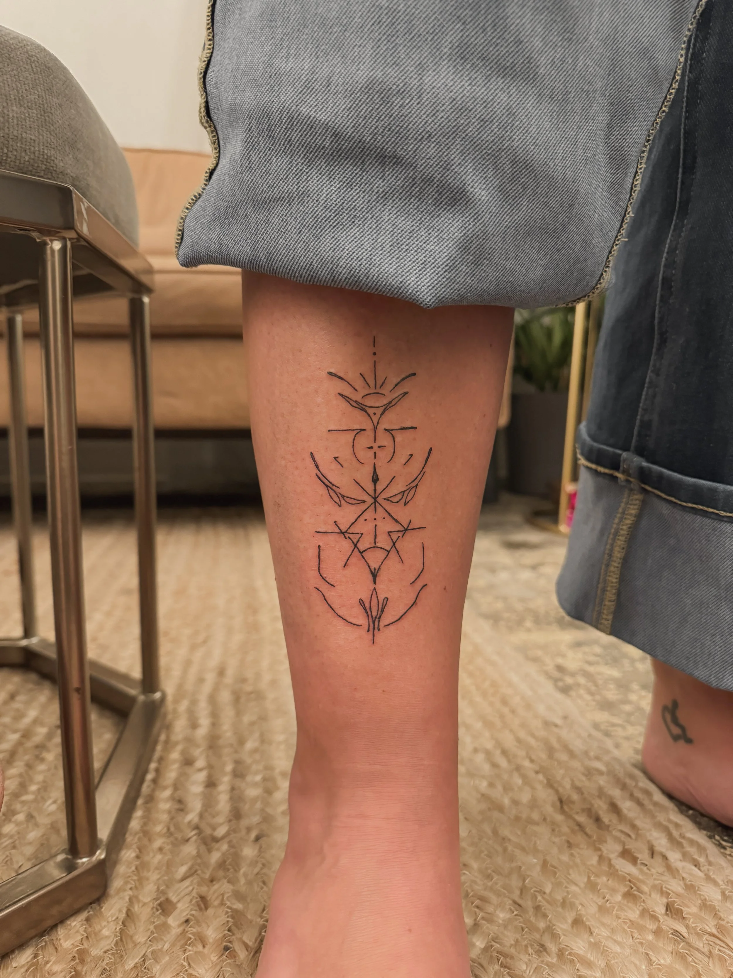 A geometric black line tattoo on a person's lower leg, featuring symmetrical shapes, lines, and abstract symbols.