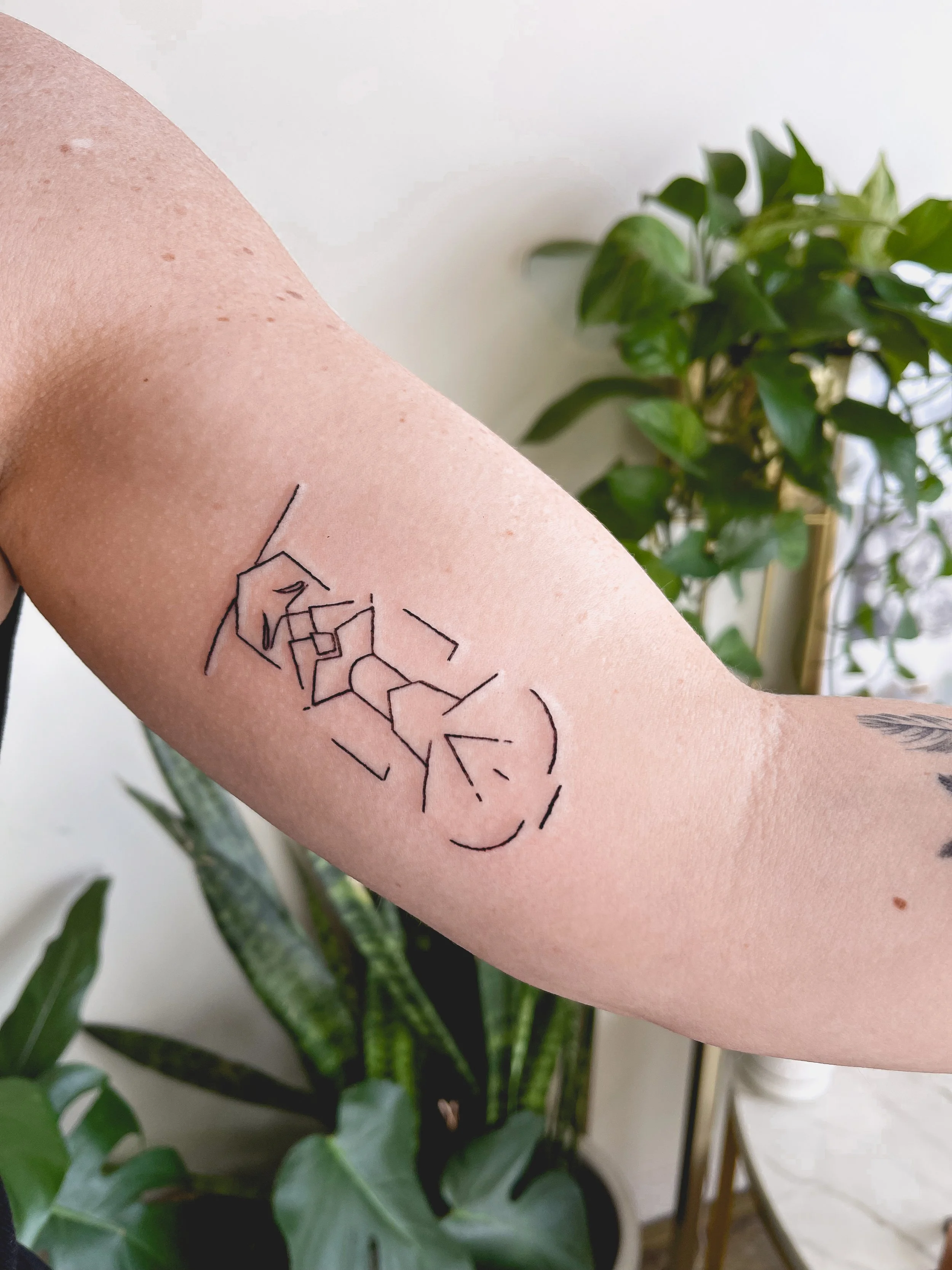 Tattoo of a line-drawn face with abstract geometric shapes on a person's forearm, with green houseplants in the background.