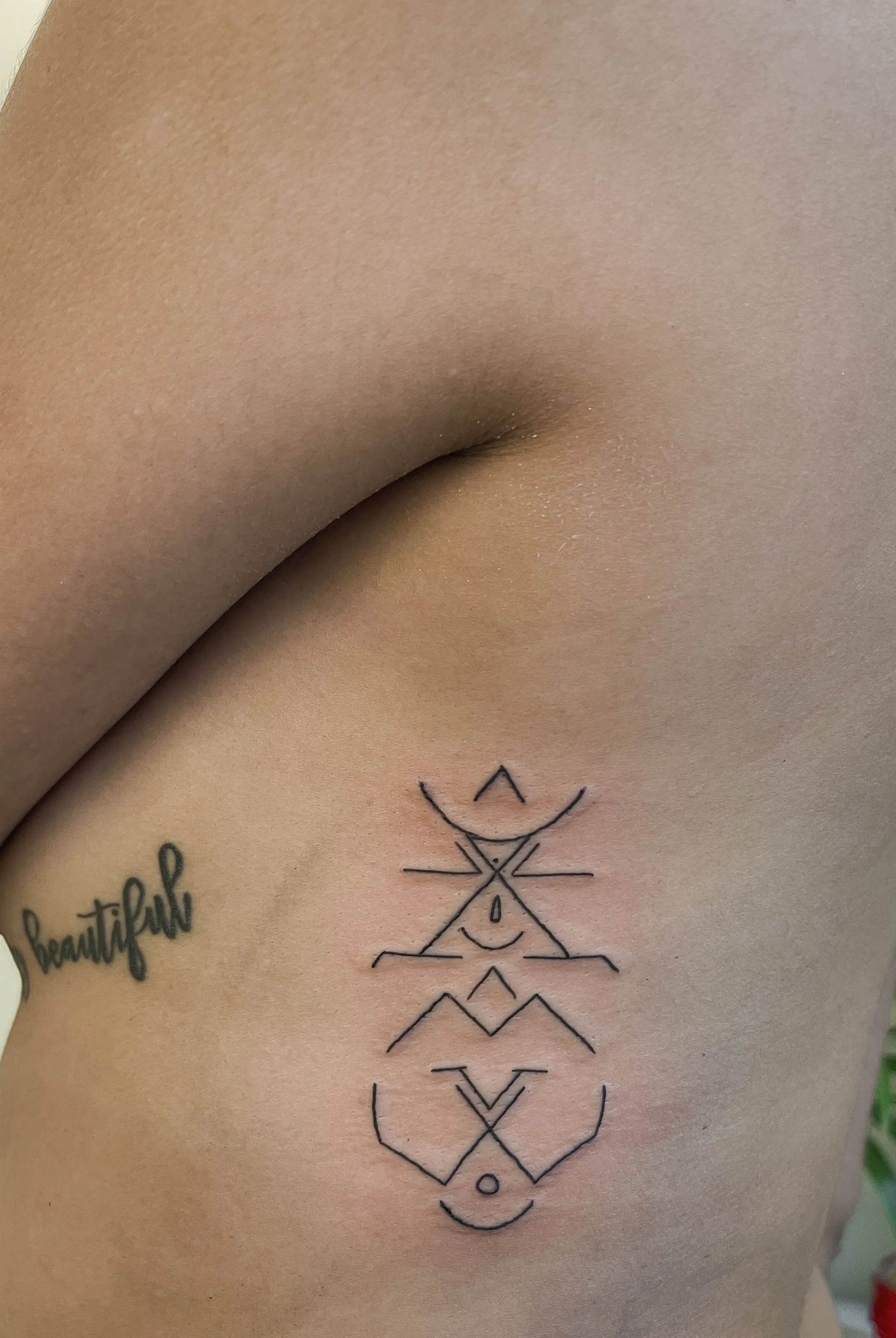 A close-up of a person's side with a geometric tattoo and a cursive tattoo that says 'beautiful'.