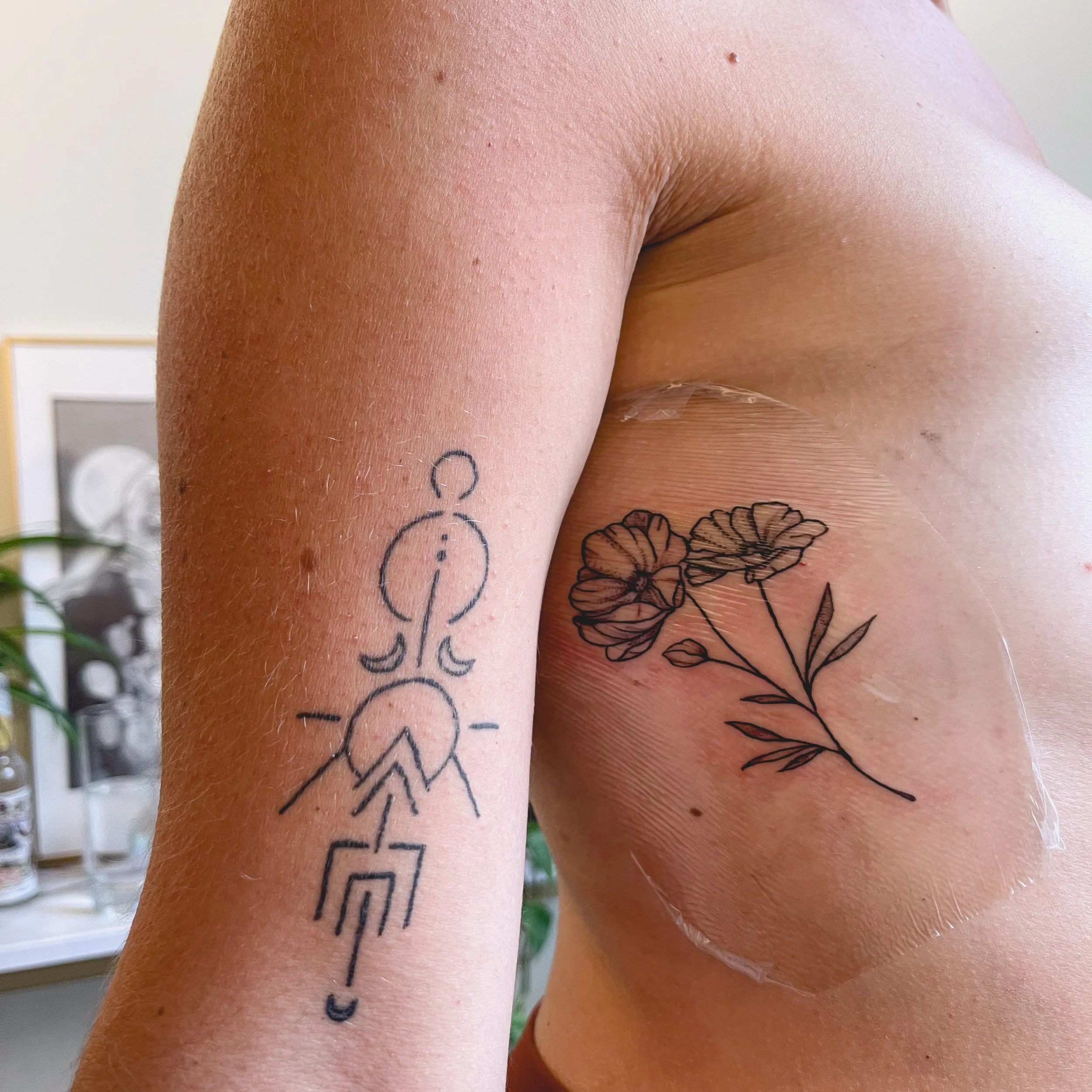 Close-up of a person's upper arm with two tattoos: a minimalistic, abstract line drawing of a person with geometric shapes on the left arm, and a simple line drawing of a flower with two blooms on the right side, partially covered with transparent pr