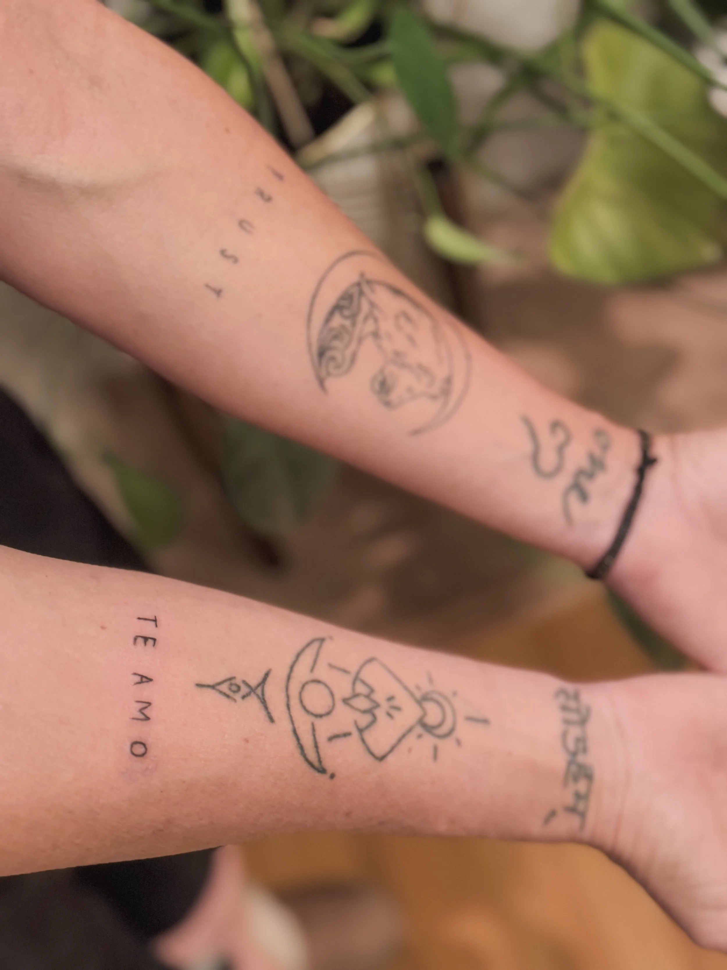 Close-up of two arms with tattoos, one with a circular tattoo featuring a mountain and moon, and another with a black bracelet and various tattoos, including a small word and symbols.