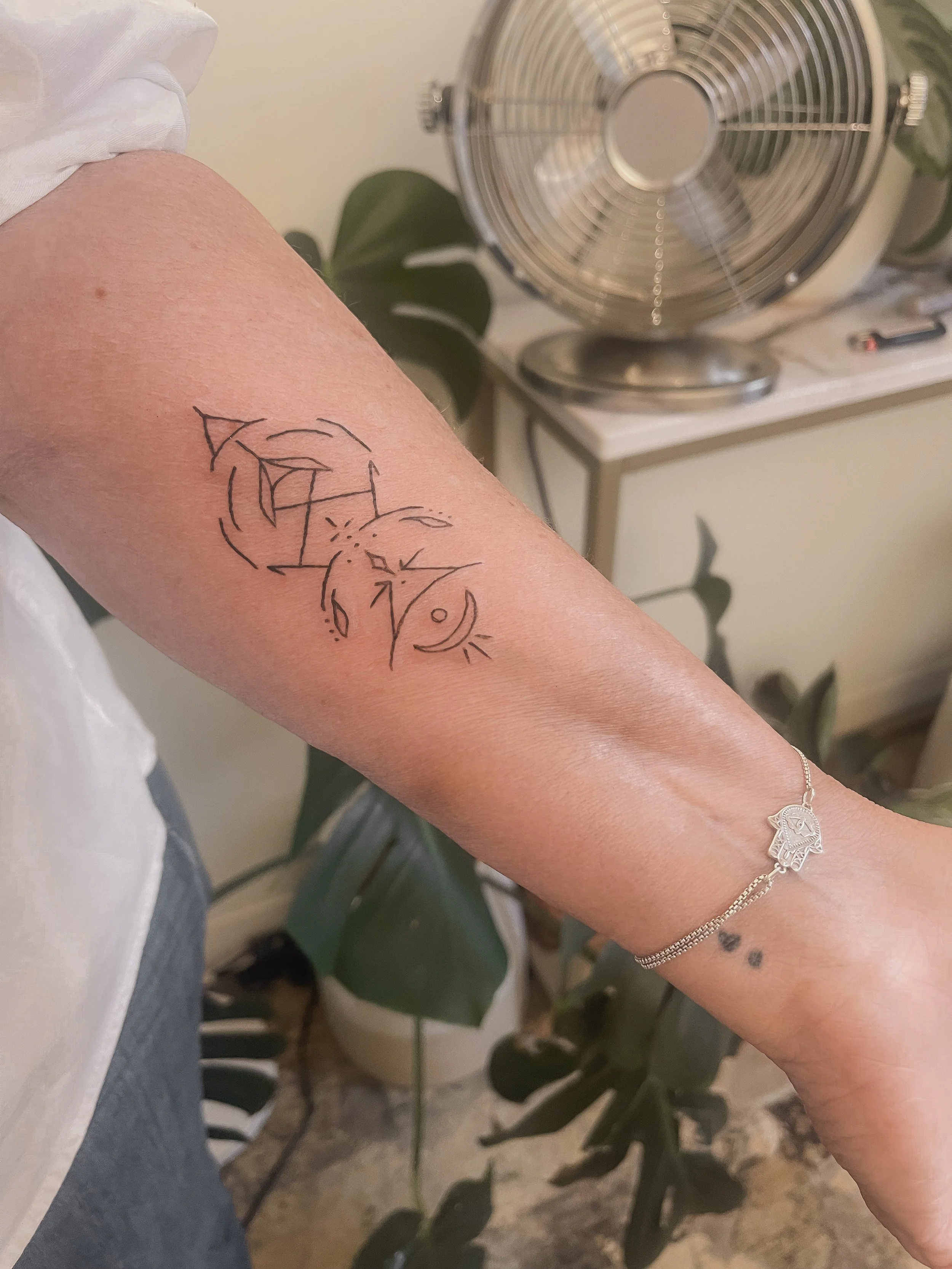 Tattoo on person's arm featuring abstract line art of faces, with a small black heart tattoo and an anklet bracelet.