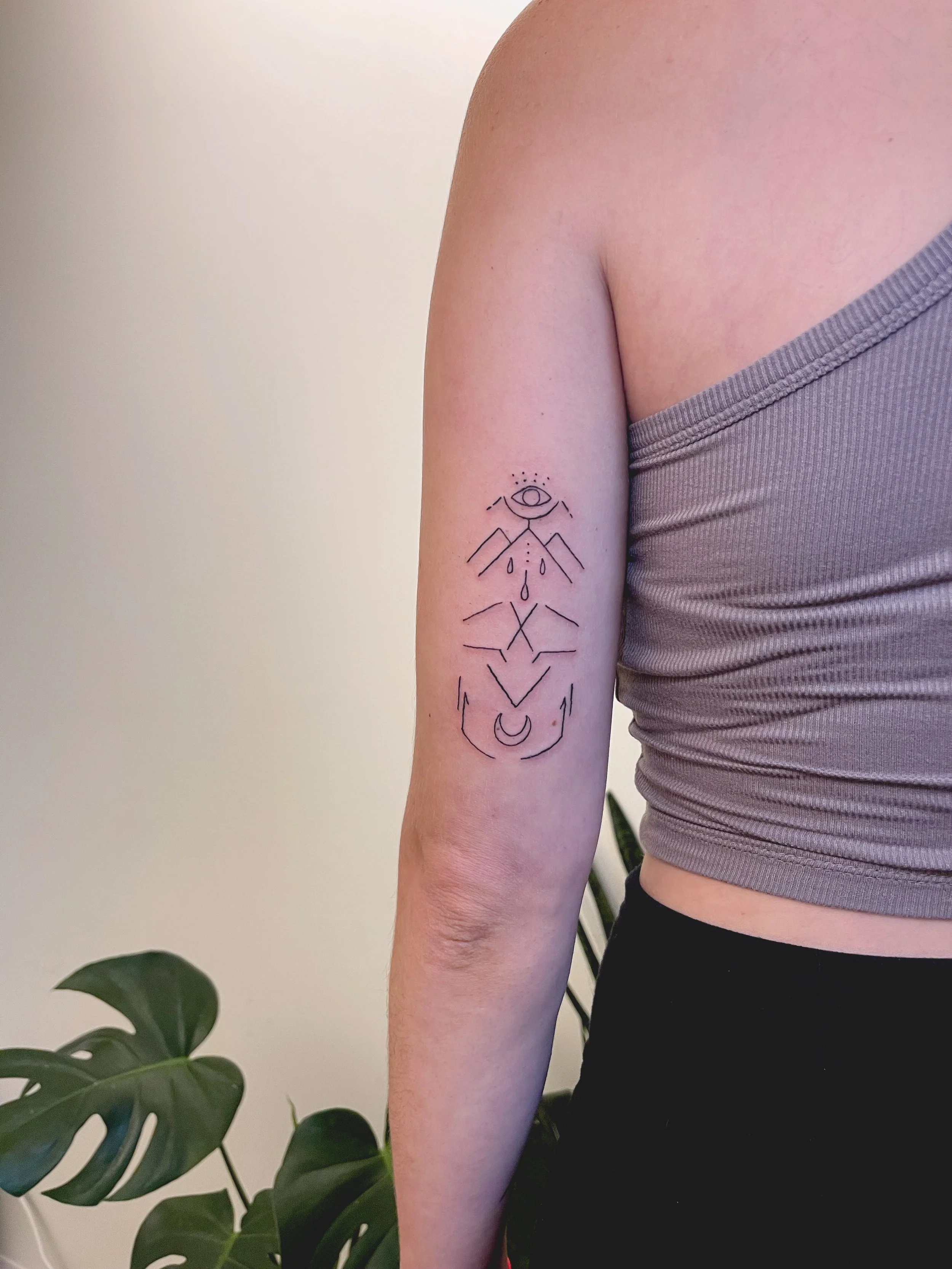 A person's upper arm with a minimalist black line tattoo featuring an eye, mountain shapes, drops, and a crescent moon.