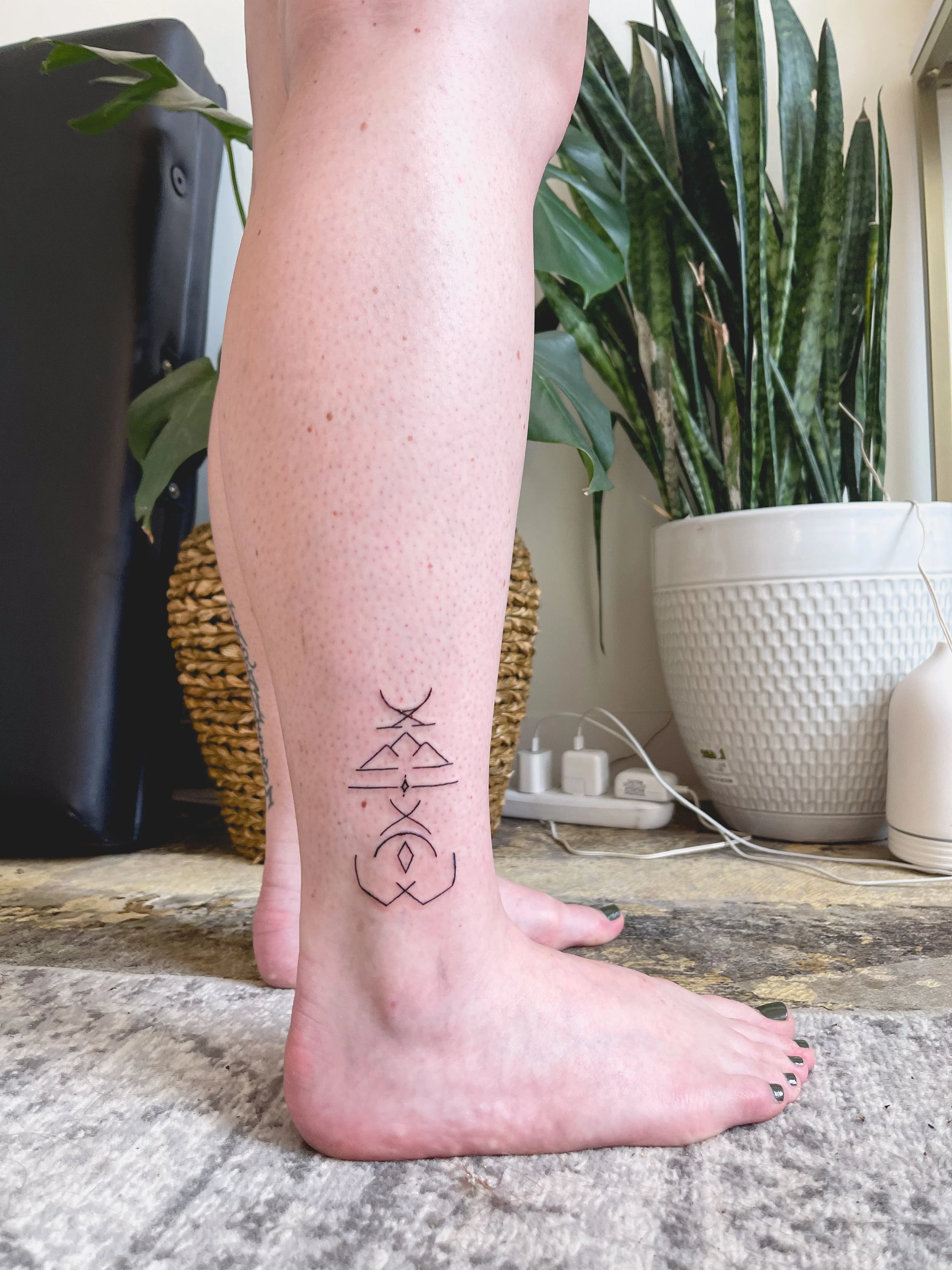 A person's lower leg and foot with a geometric tattoo on the side, next to houseplants and an electrical outlet.