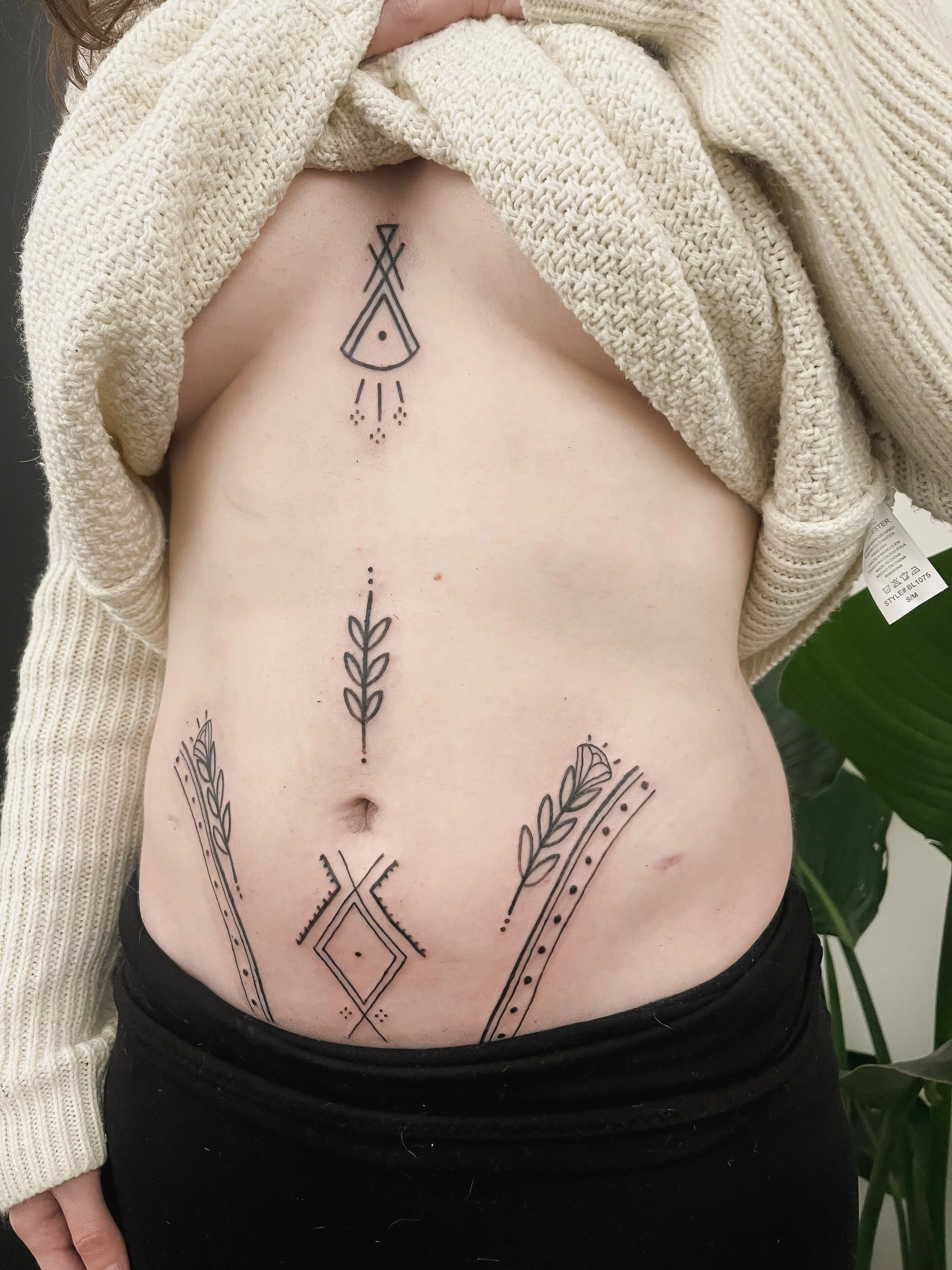 A person's lower chest and upper abdomen with four black geometric tattoos, with beige knit and wool sweaters seen at the top and black pants at the bottom.