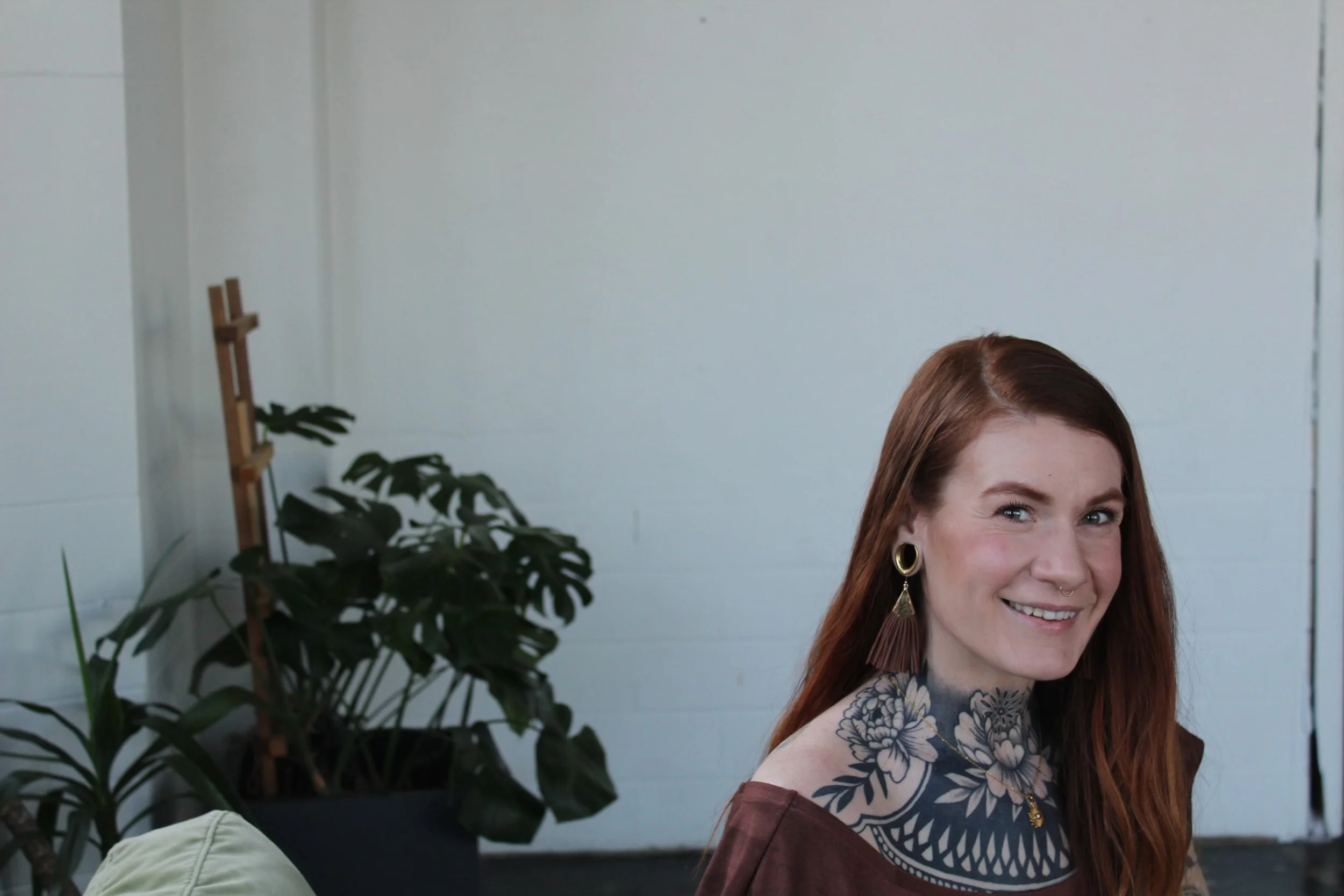 A woman with long red hair and tattoos on her neck and chest, smiling at the camera, sitting indoors with a potted plant and a white wall in the background.
