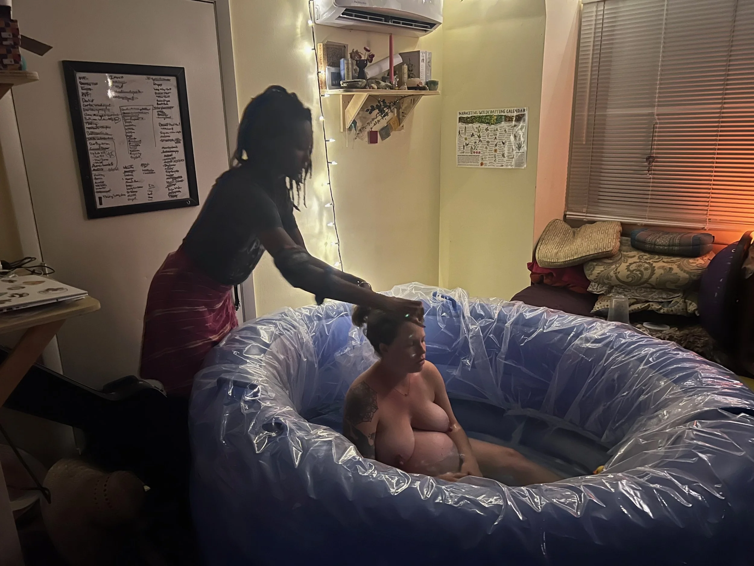 A woman sitting in an inflatable pool with her doula supporting her in labor, in a dimly lit room with shelves and cushions.