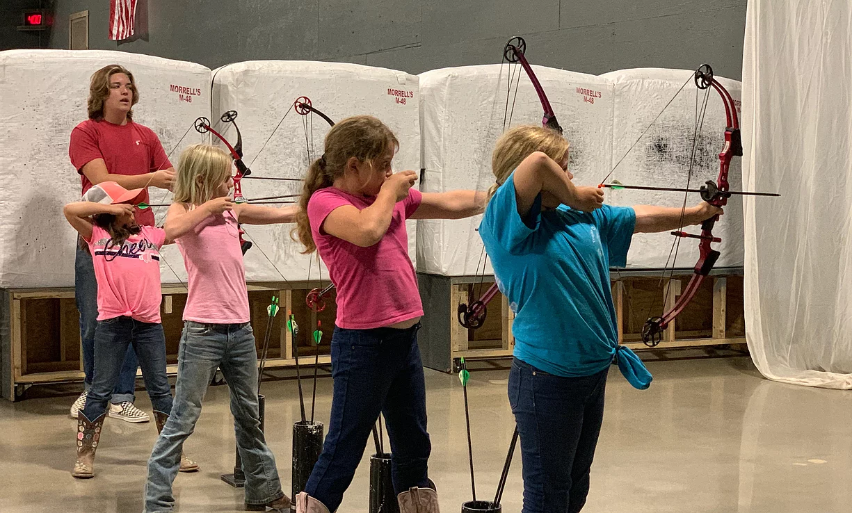 Four girls and one woman practicing archery indoors, with targets and padded walls in the background.
