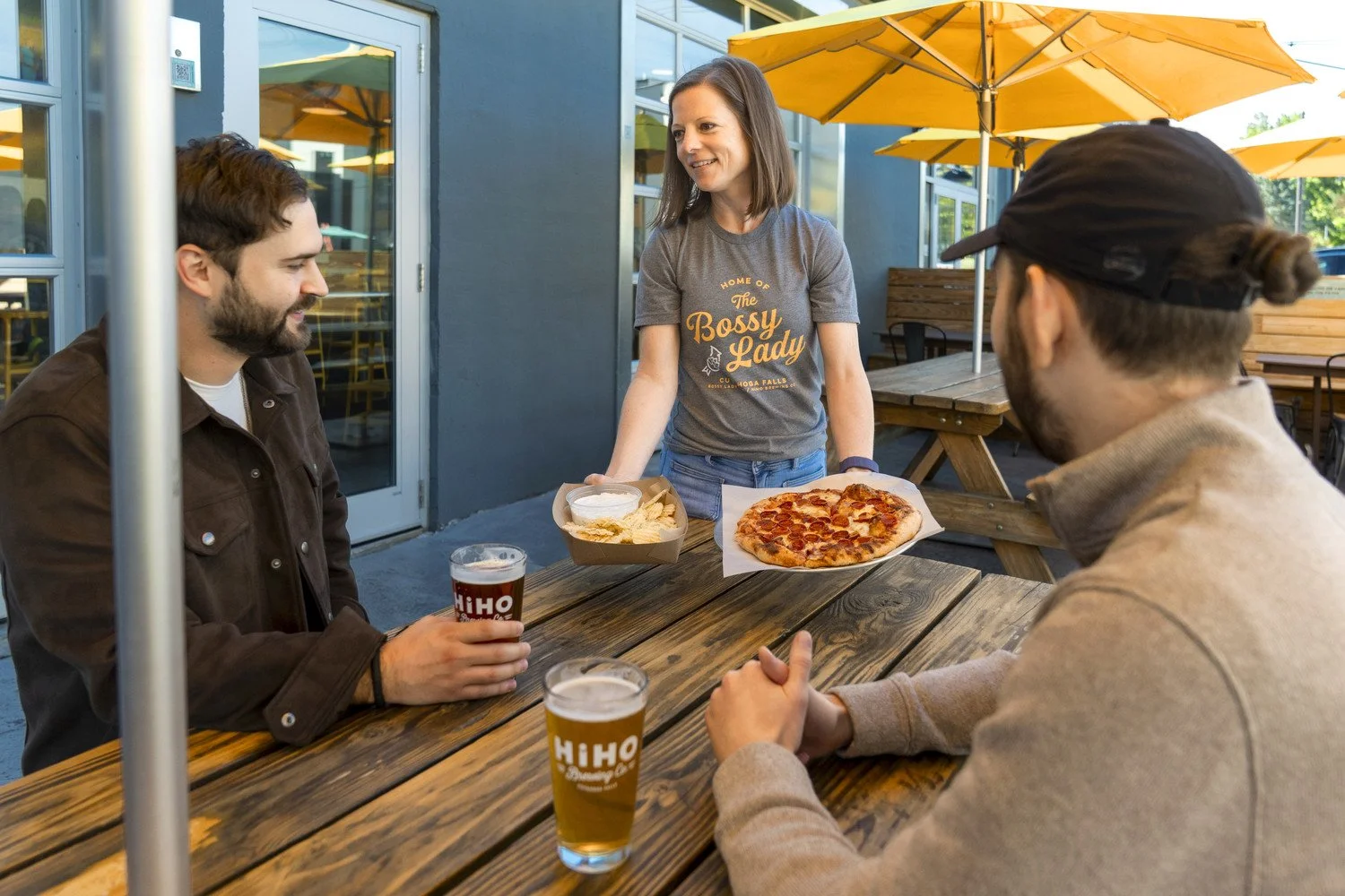 A woman serving pizza and chips to two men at an outdoor table with drinks, under yellow umbrellas, near a modern building.