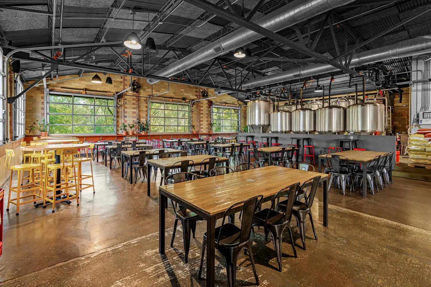 Interior of HiHO Brewing Co. with wooden tables, black and yellow chairs, large windows, and brewing tanks in the background.