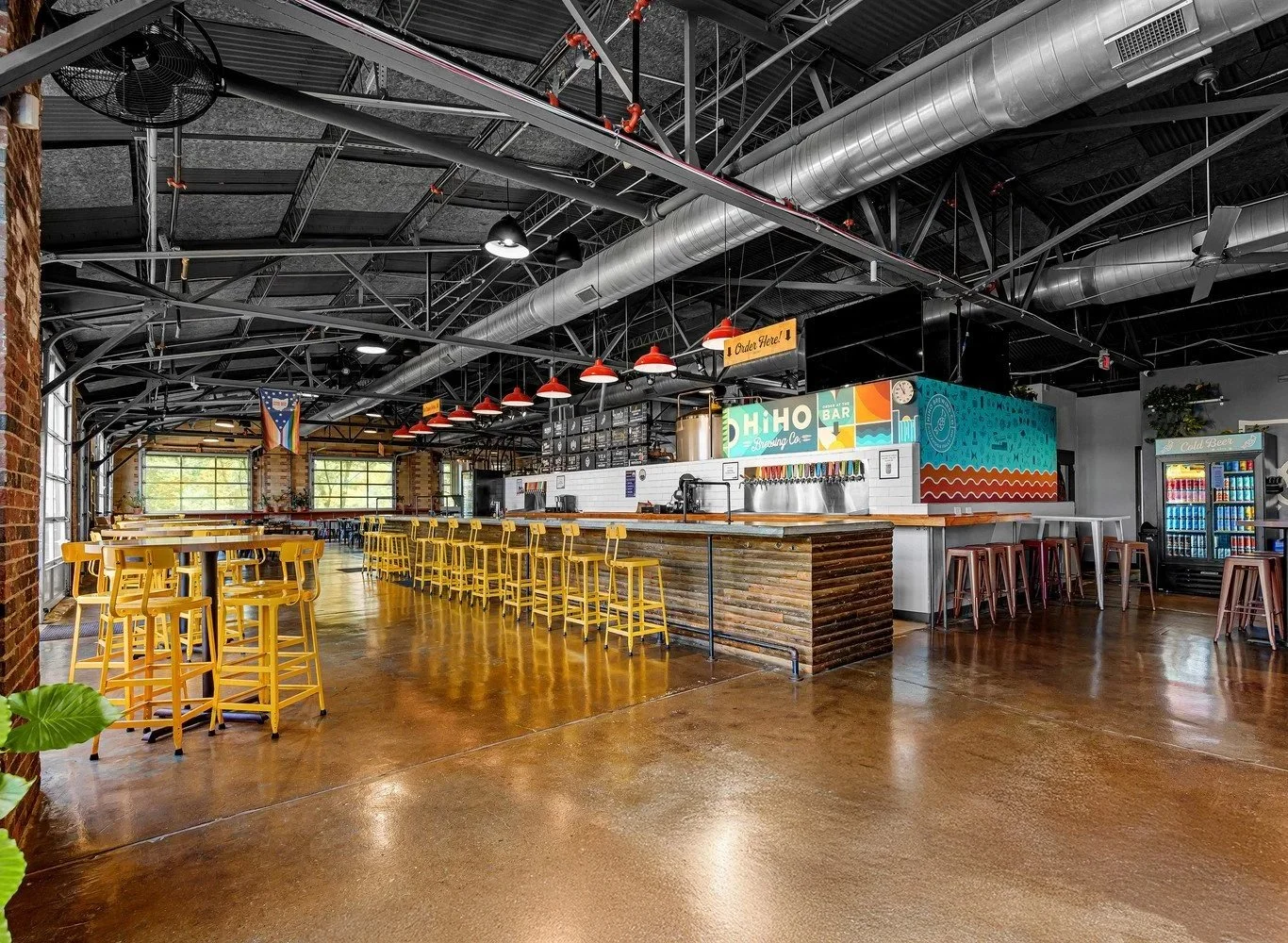 HiHO Brewing Co., brightly lit with yellow high stools at the bar and yellow chairs around tables, with industrial ceiling elements and a colorful mural on the back wall.