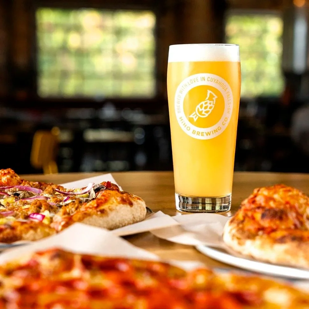 A pint of HiHO Brewing Co. beer on a table, surrounded by pizza slices, against a background of a cozy indoor setting with windows showing greenery outside.
