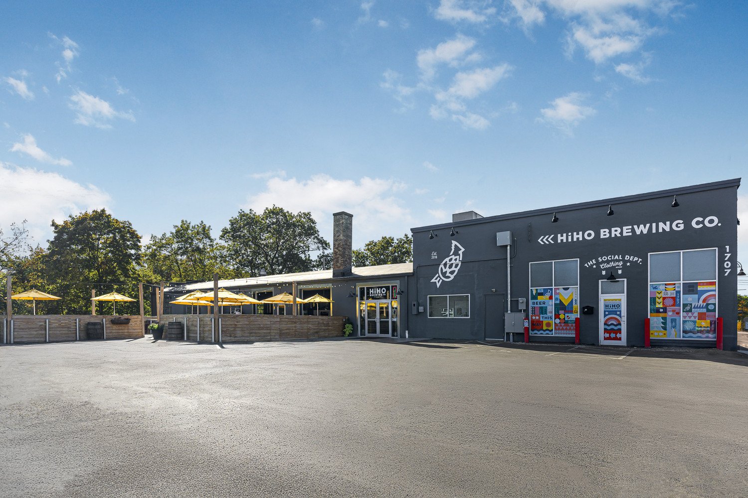 Exterior of HiHO Brewing Co. building with colorful murals on the front, outdoor seating with yellow umbrellas, and a spacious parking area under a blue sky.
