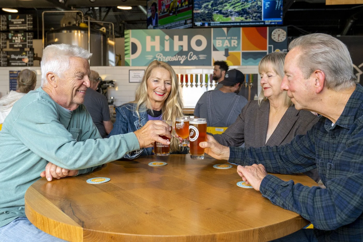 Four adults sitting around a wooden table at a HiHO Brewing Co., raising glasses of beer for a toast and smiling, with a brewery signage and taps visible in the background.