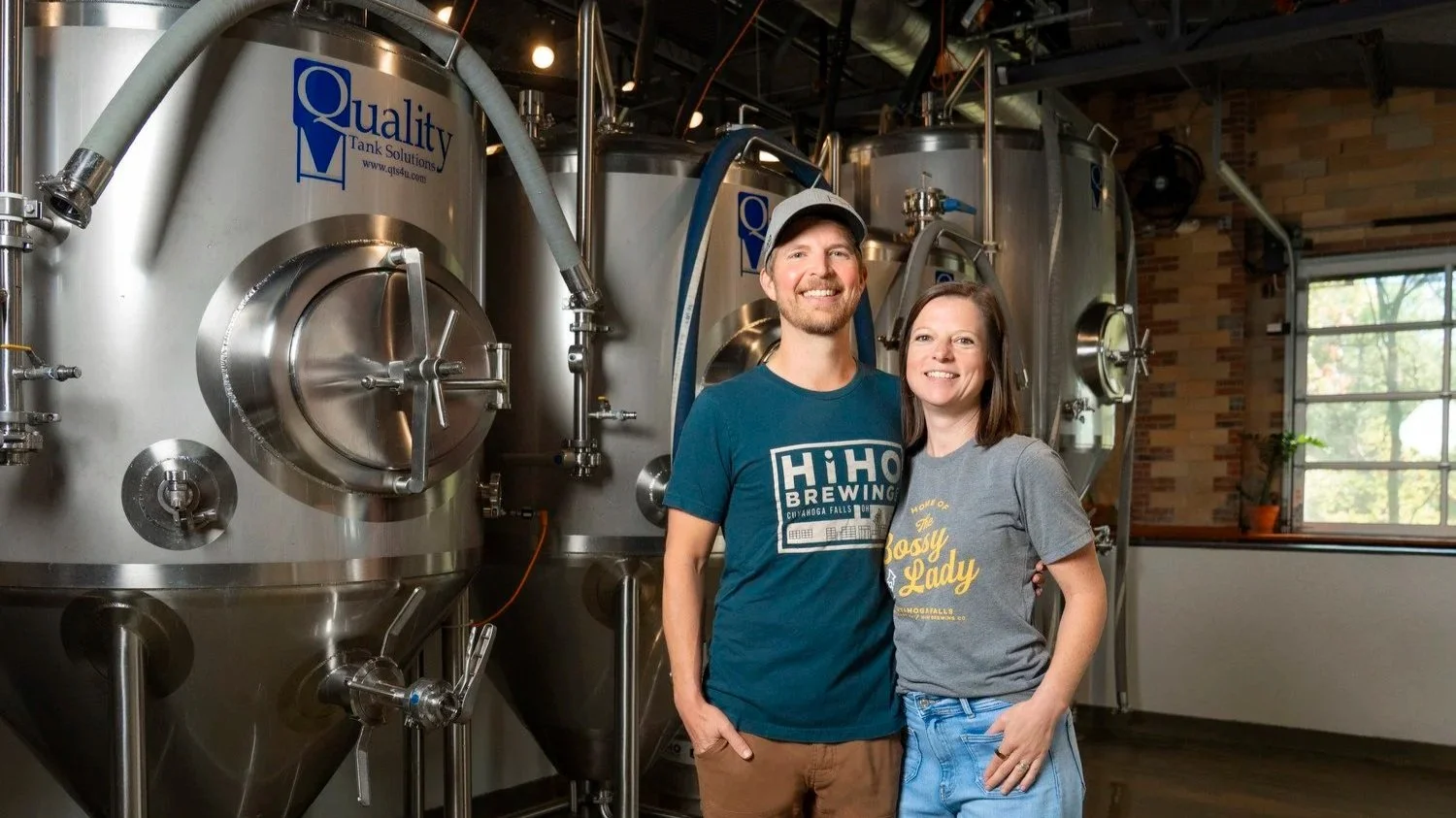 The owners of HiHO Brewing Co., Jon and Ali Hovan, standing inside a HiHO with large stainless steel brewing tanks in the background.