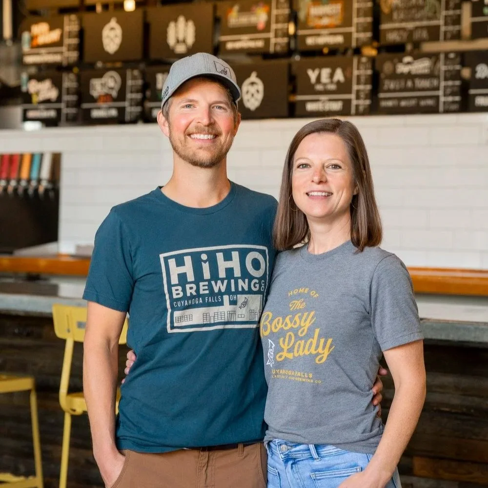 The owners of HiHO Brewing Co., Jon and Ali Hovan, both wearing casual t-shirts with HiHO logos and a tap list visible in the background.
