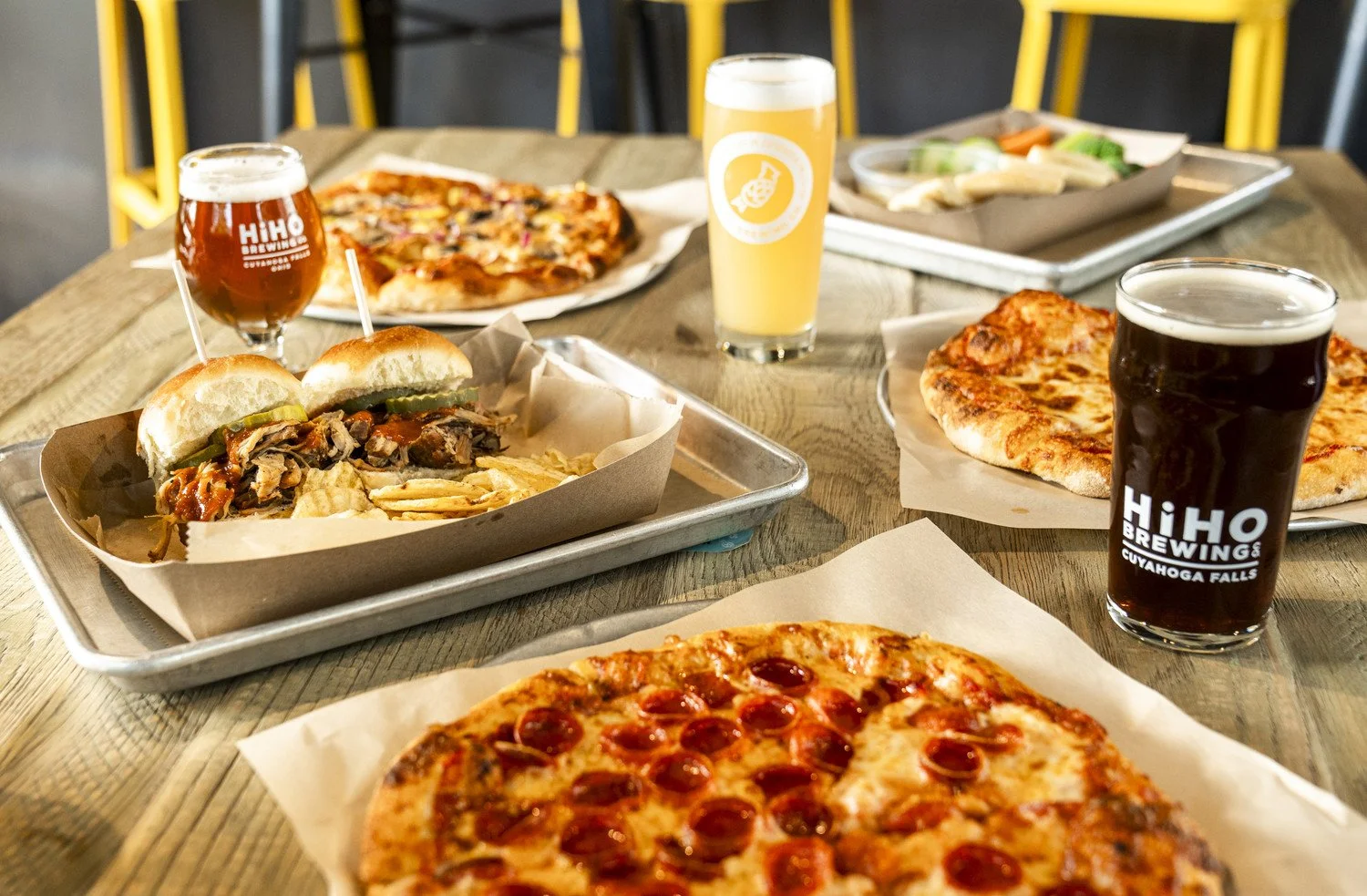 Food and drinks on a wooden table, including two pizzas, a sandwich with pickles, two glasses of dark beer, and a light-colored beverage in a glass, with some side dishes in the background.