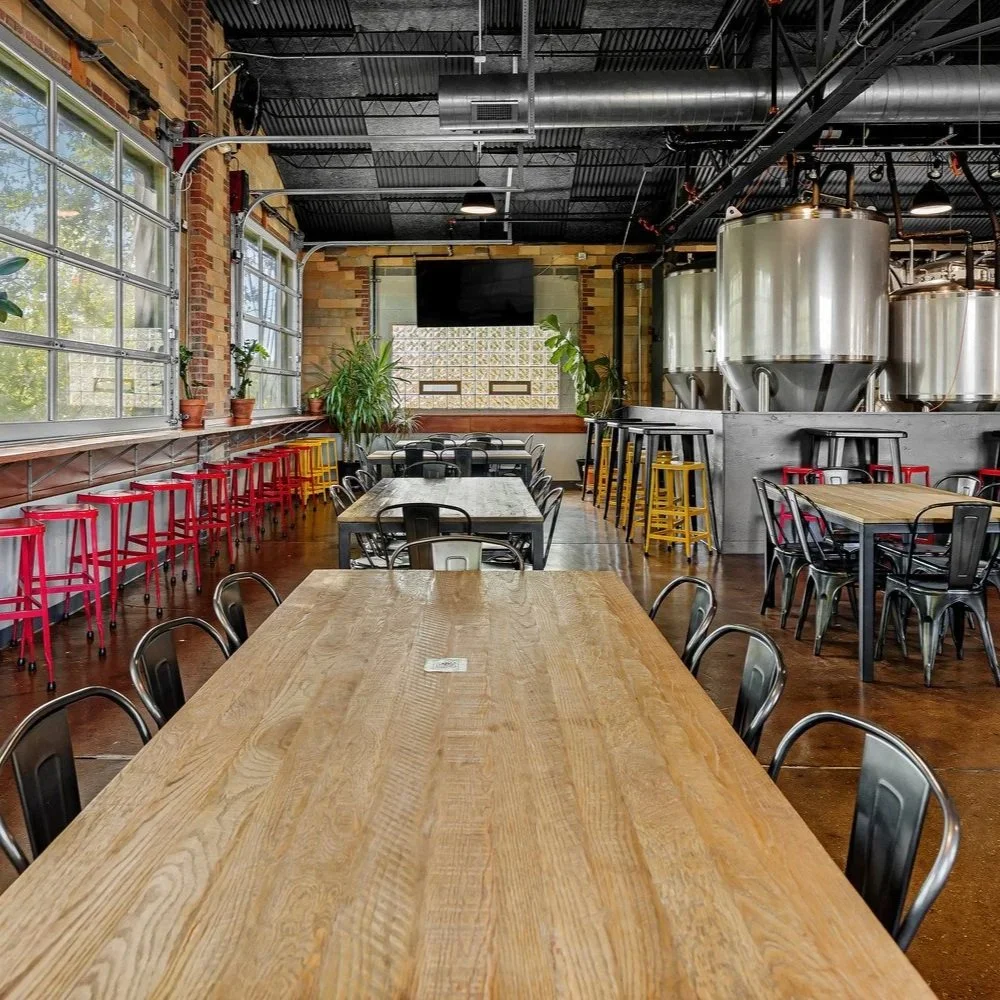 Inside of HiHO Brewing Co. with large brewing tanks, wooden tables, metal chairs, and colorful bar stools near windows.