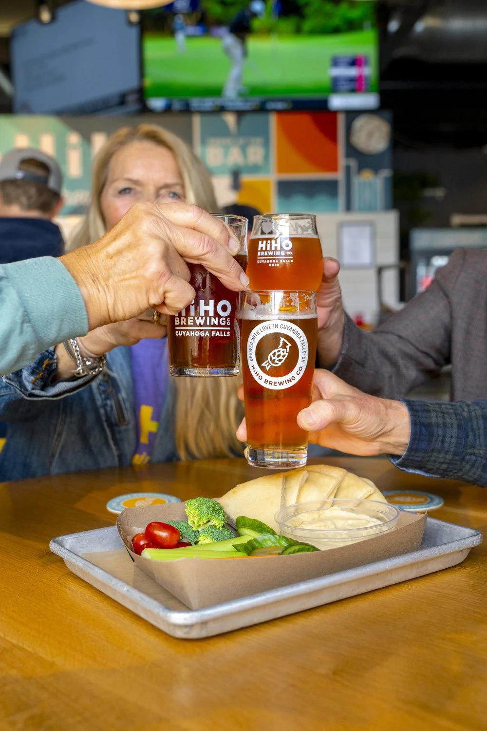 Three people clinking glasses of beer at HiHO Brewing Co., with a plate of vegetables and bread on the table in the foreground.